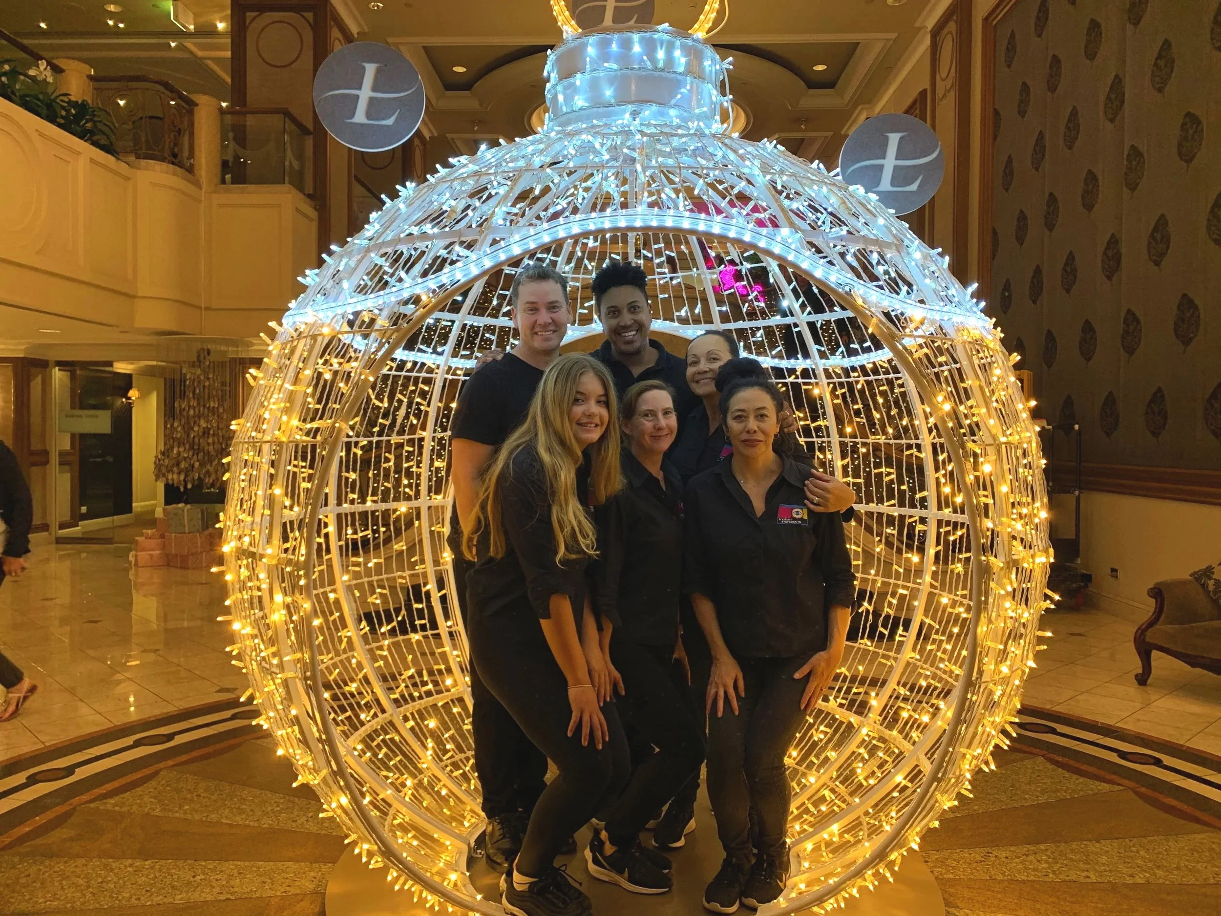 The Visual Statements Team - Group of six people, four women and two men, standing inside a glowing Christmas bauble lit up in the Langham Hotel lobby