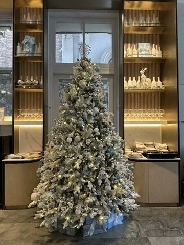 A decorated Christmas tree at Lanson Place Melbourne with white and silver ornaments, lights, and wrapped presents underneath, placed in front of a window with built-in shelves containing decorative items.
