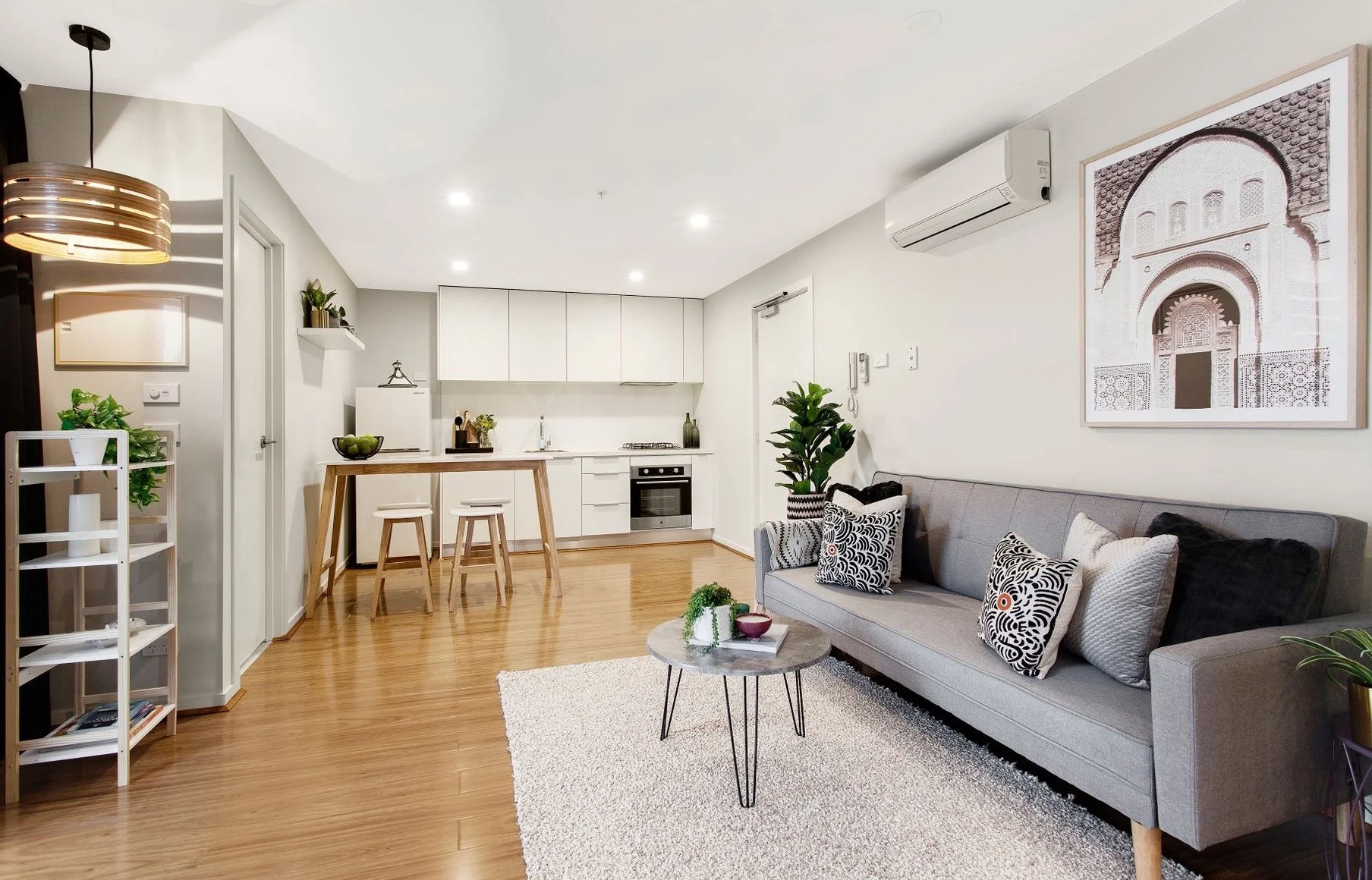 Living room and kitchen area with wood flooring, a gray sofa with black and white patterned pillows, a small round coffee table with decorative items, and a white rug. The kitchen features white cabinets, a white refrigerator, and a small wooden dini