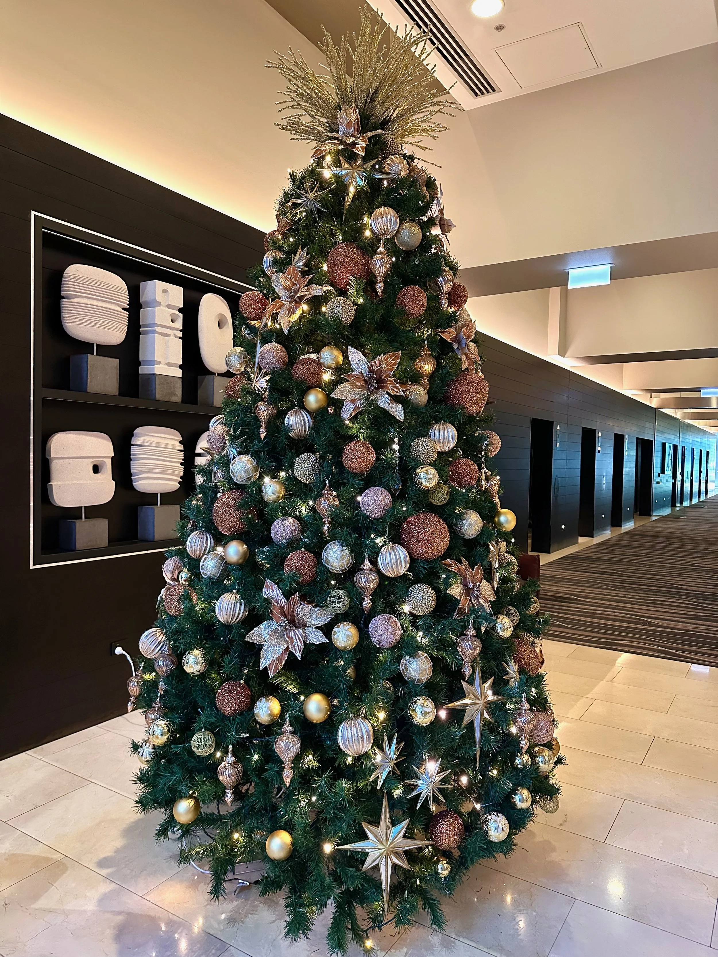 A decorated Christmas tree with gold, bronze, and silver ornaments, including star and leaf shapes, topped with branches, situated in an indoor setting with modern decor, next to a wall with abstract sculptures.