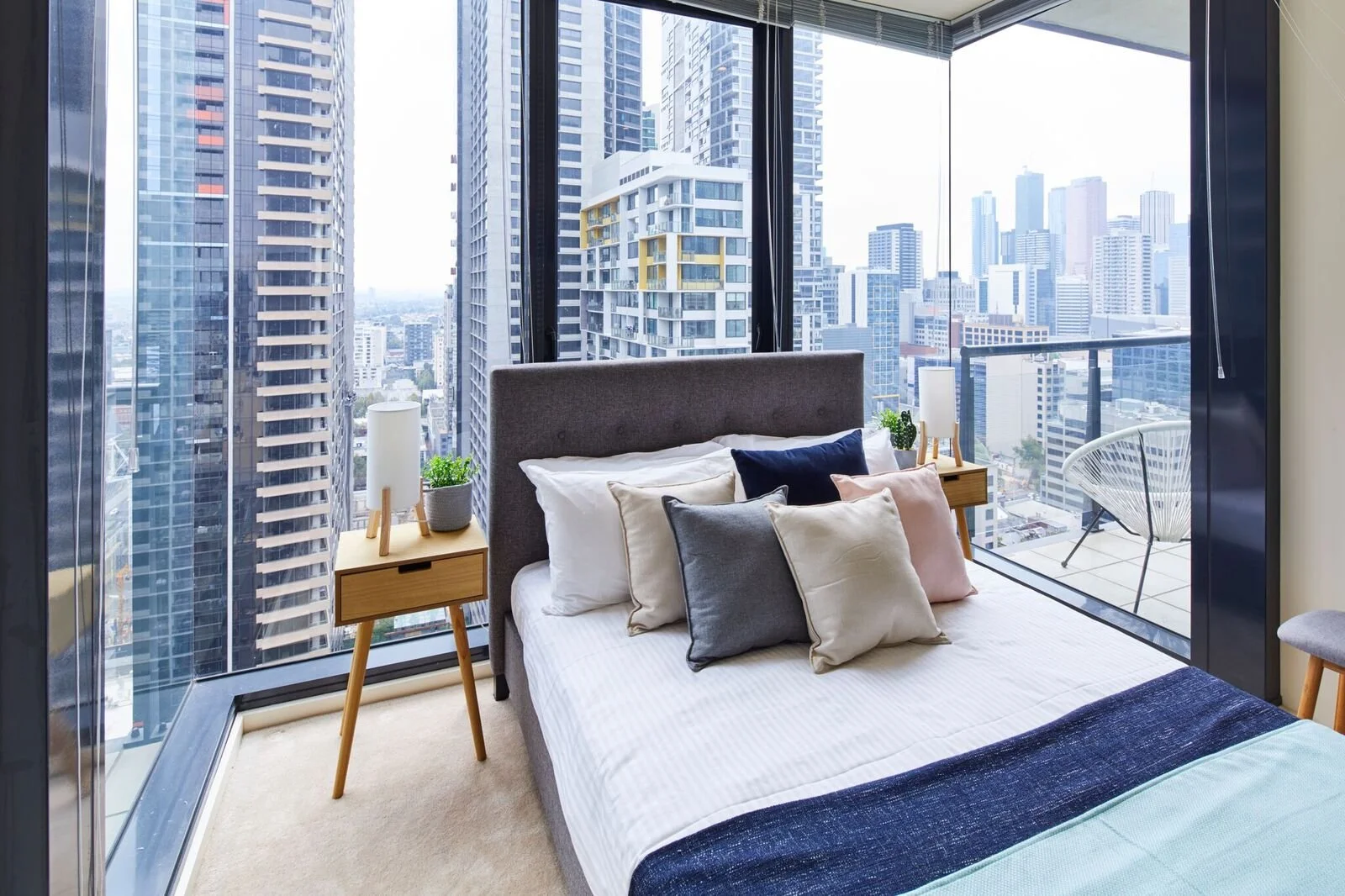 Modern bedroom with a large bed and multiple pillows, floor-to-ceiling windows showing a city skyline with tall buildings, nightstand with a lamp and plants on each side of the bed, and a balcony with outdoor chair.