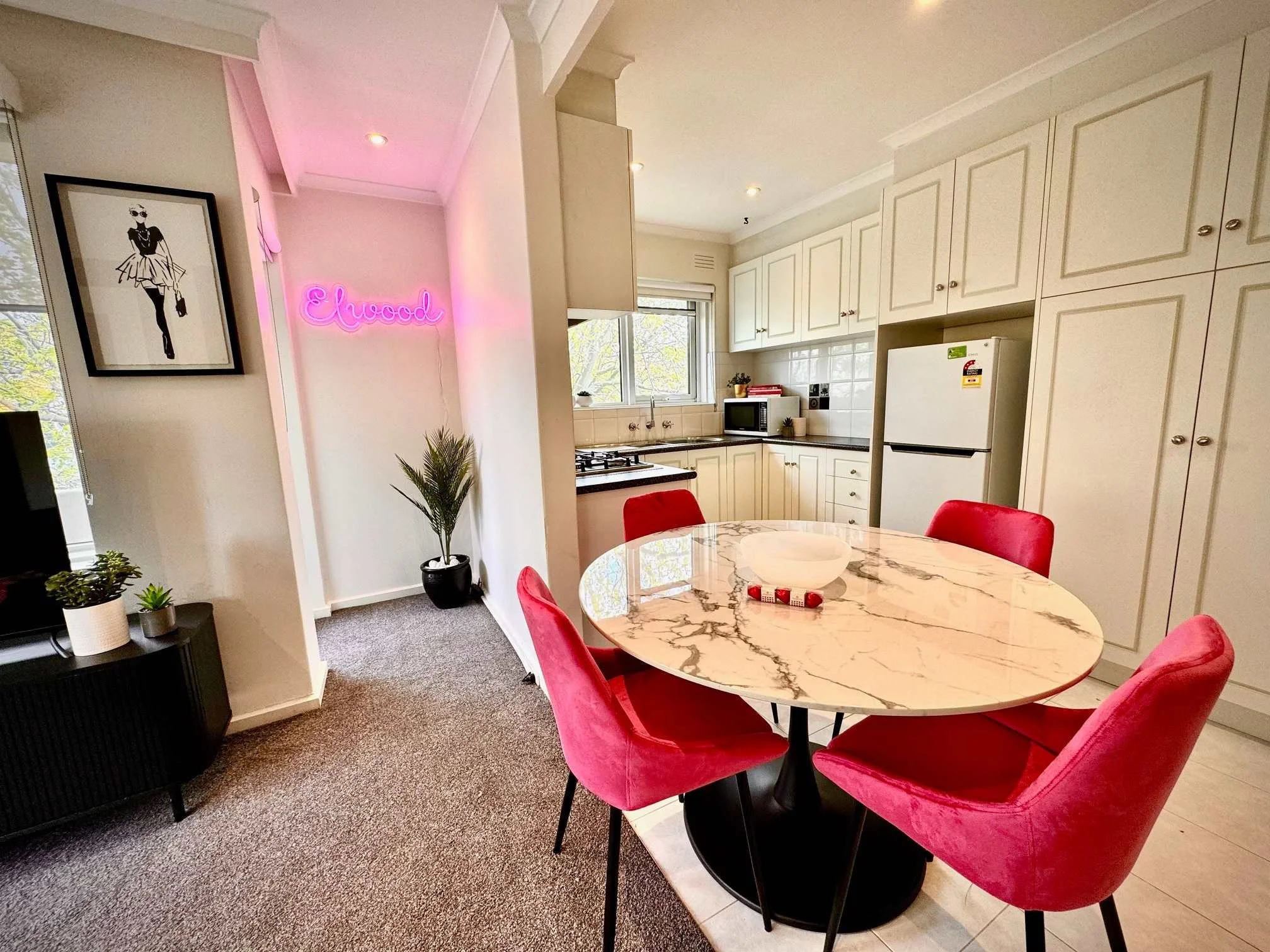 A kitchen and dining area with white cabinetry, a round marble table with red velvet chairs, a potted plant, and a neon pink sign that says "Enood" on the wall. There are kitchen appliances and a window with a view of trees outside.