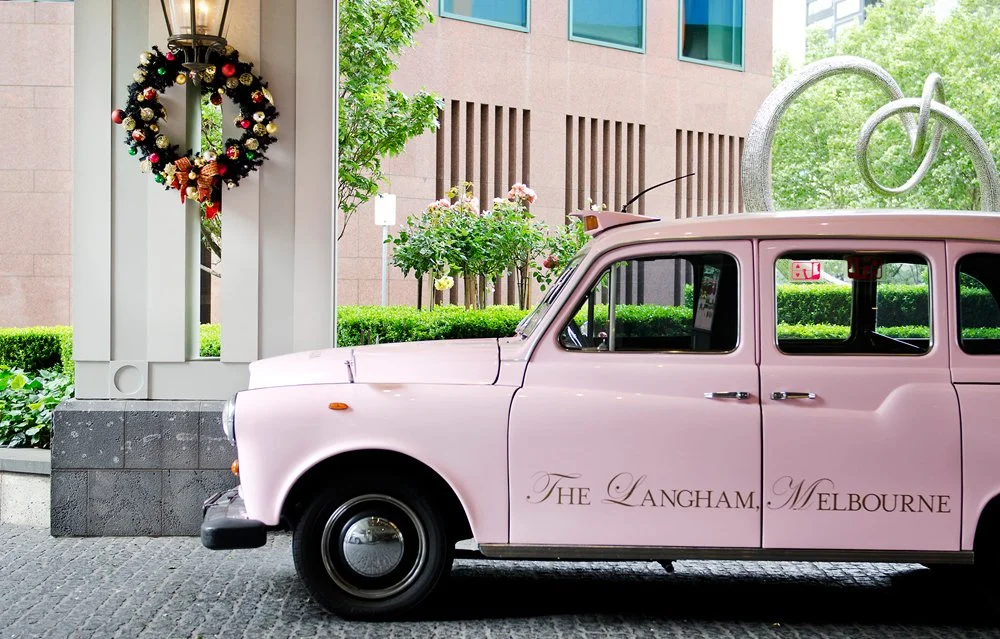 A vintage pink taxi cab with words "The Langham, Melbourne" on the door, parked next to a building decorated with a wreath for Christmas or holiday season, with green trees and a modern building in the background.