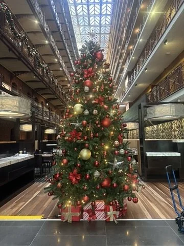 Large decorated Christmas tree at the Intercontinental Hotel Melbourne with red and gold ornaments, ribbons, and lights in a hotel lobby with multiple floors, glass ceiling, and gift boxes at the base.