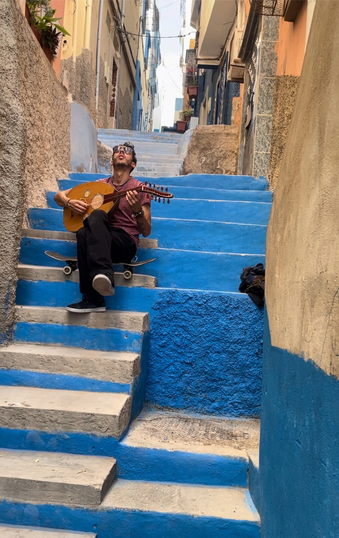 A man sitting on colorful stairs playing a stringed instrument in an alleyway with buildings on either side.