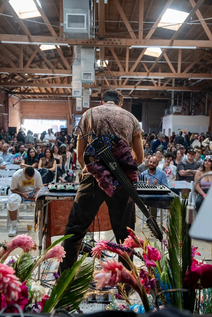 A performer with a guitar on his back stands on stage in front of a large audience in a spacious hall with wooden beams and skylights, surrounded by pink flowers in the foreground.