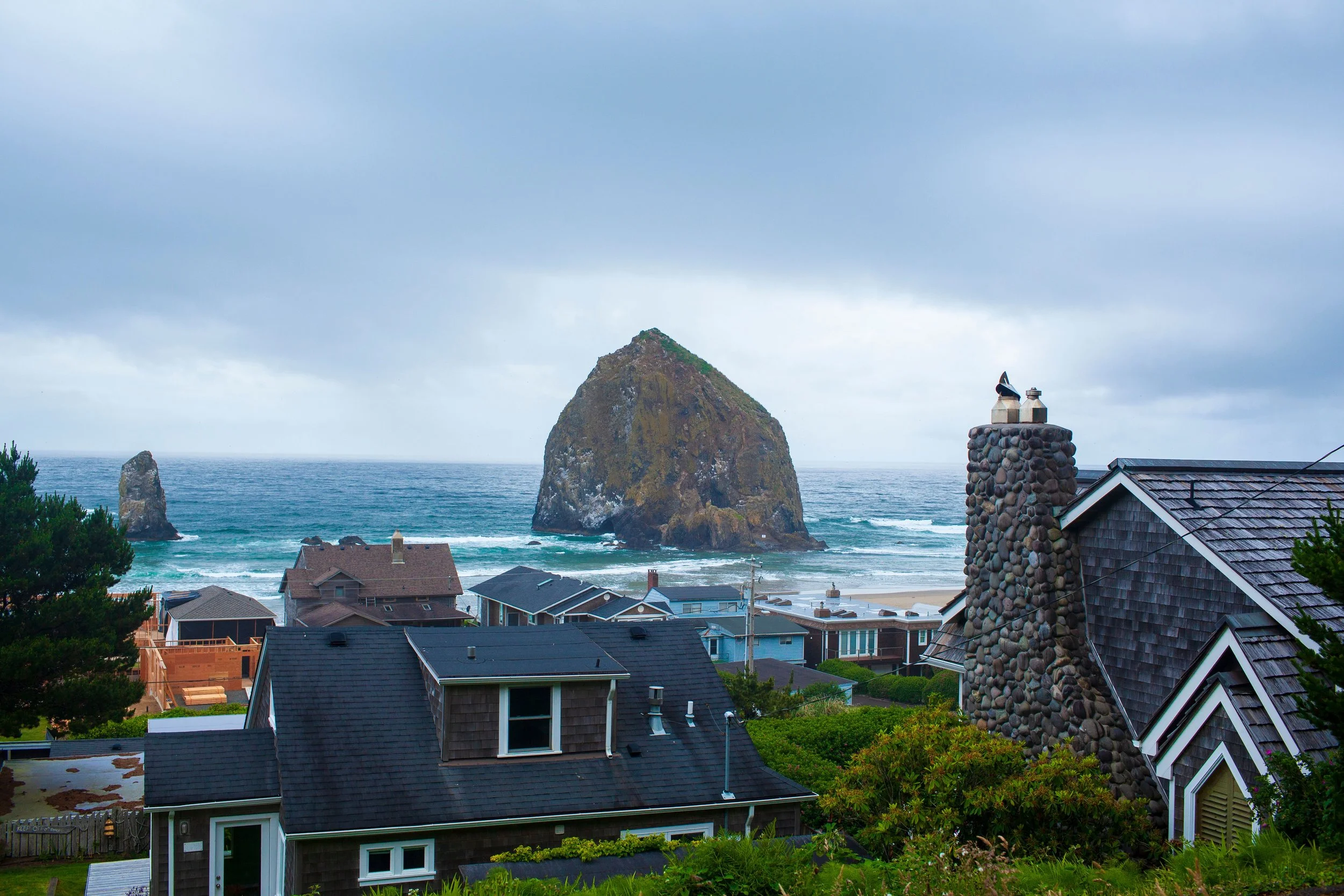 Coastal view with houses in foreground, large sea stacks in the ocean, and a cloudy sky above.