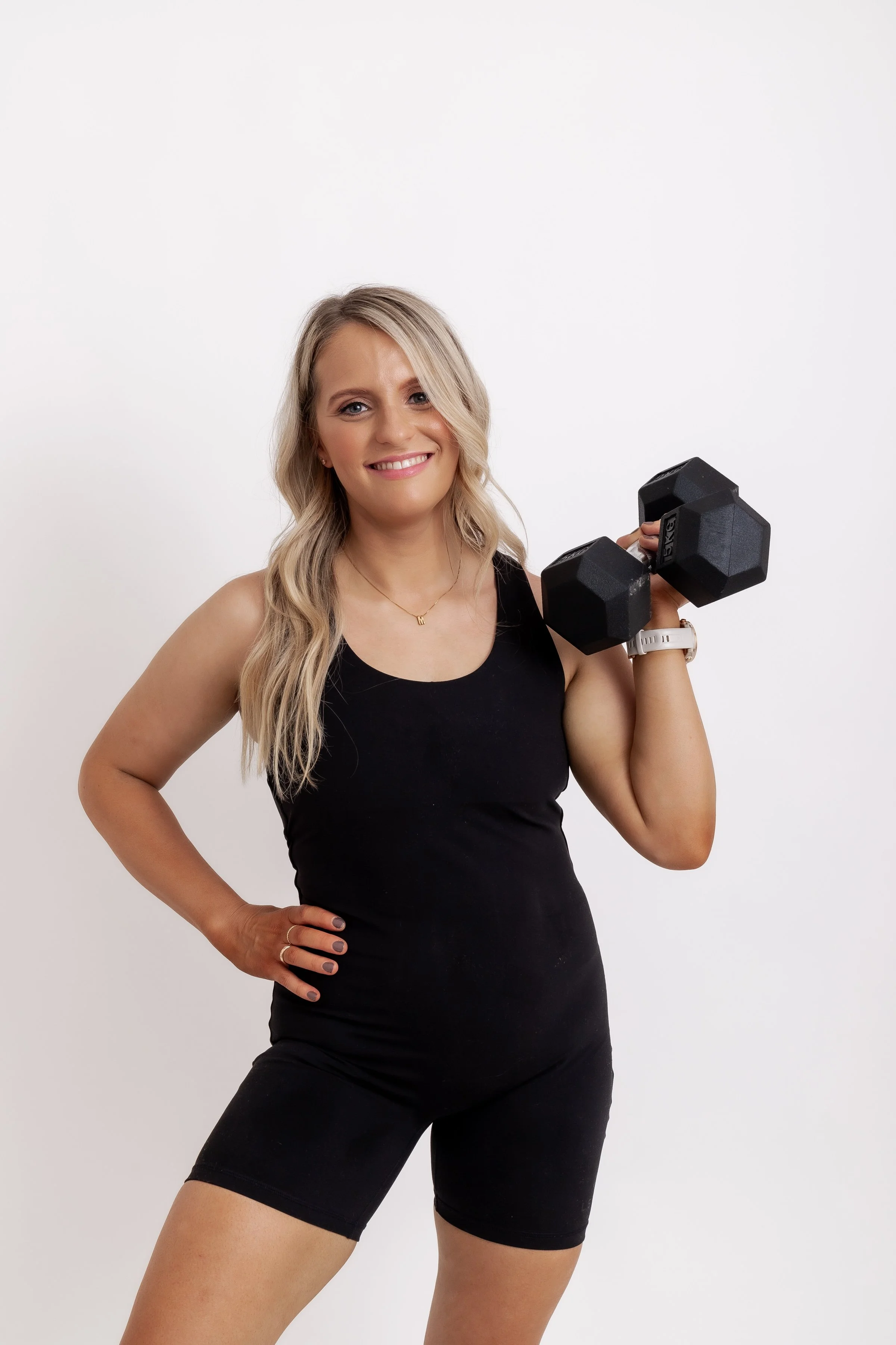 A smiling woman in black athletic wear holding a dumbbell in her right hand, standing against a plain white background.
