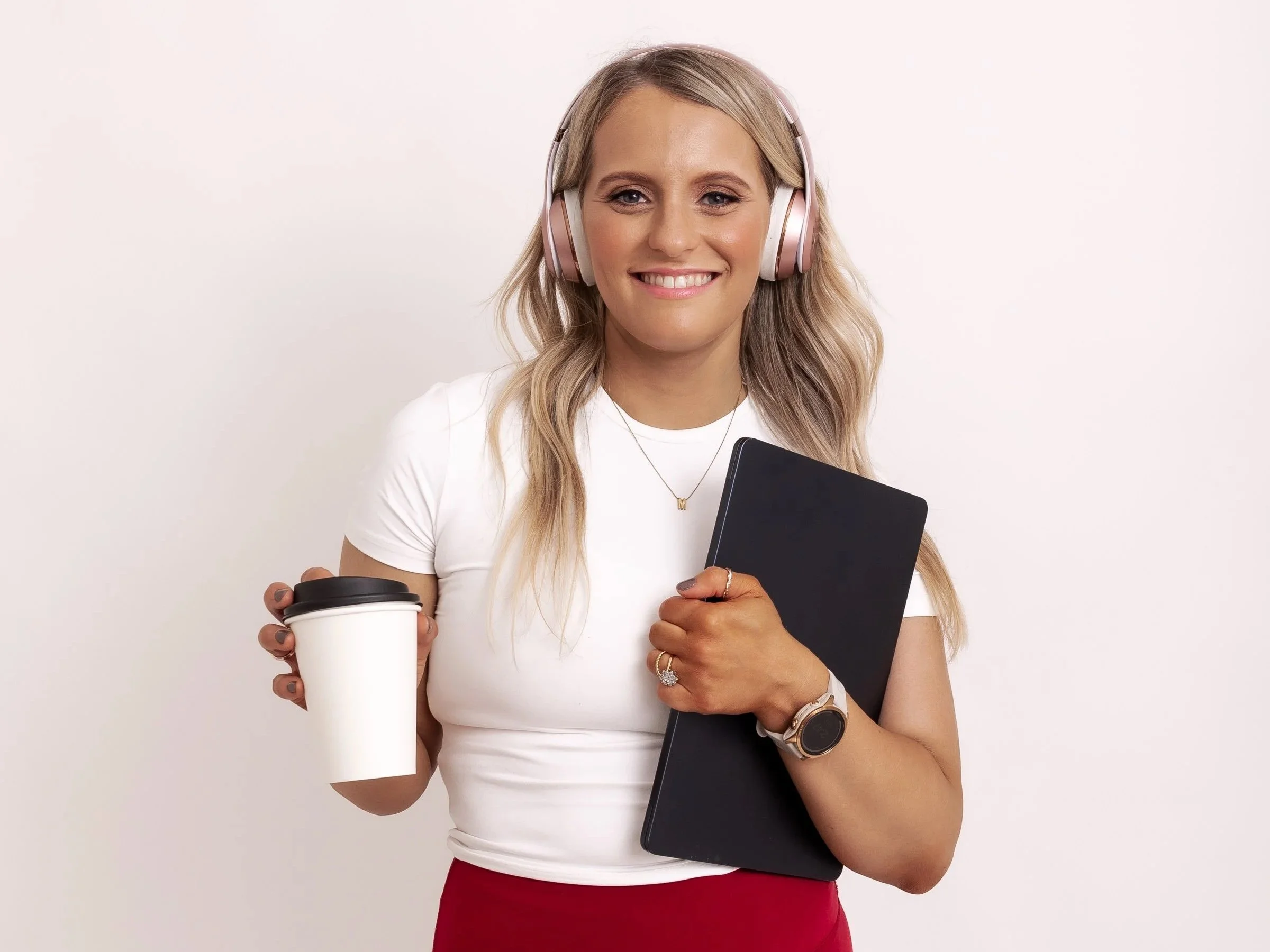 A smiling woman with blonde hair wearing pink headphones, holding a black tablet in her left arm and a white to-go coffee cup in her right hand, standing against a plain white background.