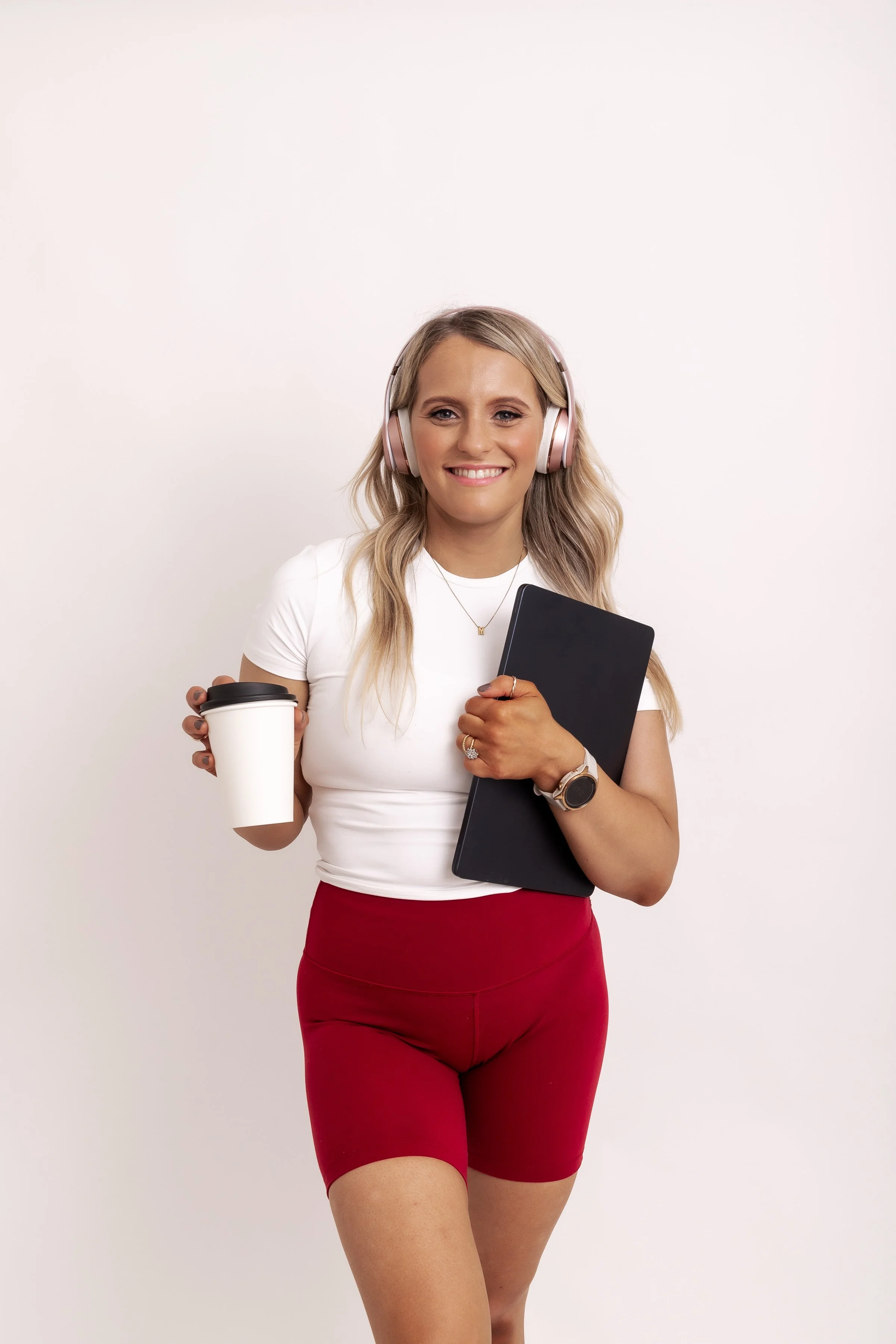 A woman with long blonde hair wearing a white t-shirt, red shorts, and pink headphones, smiling and holding a black notebook and a white coffee cup in front of a plain white background.