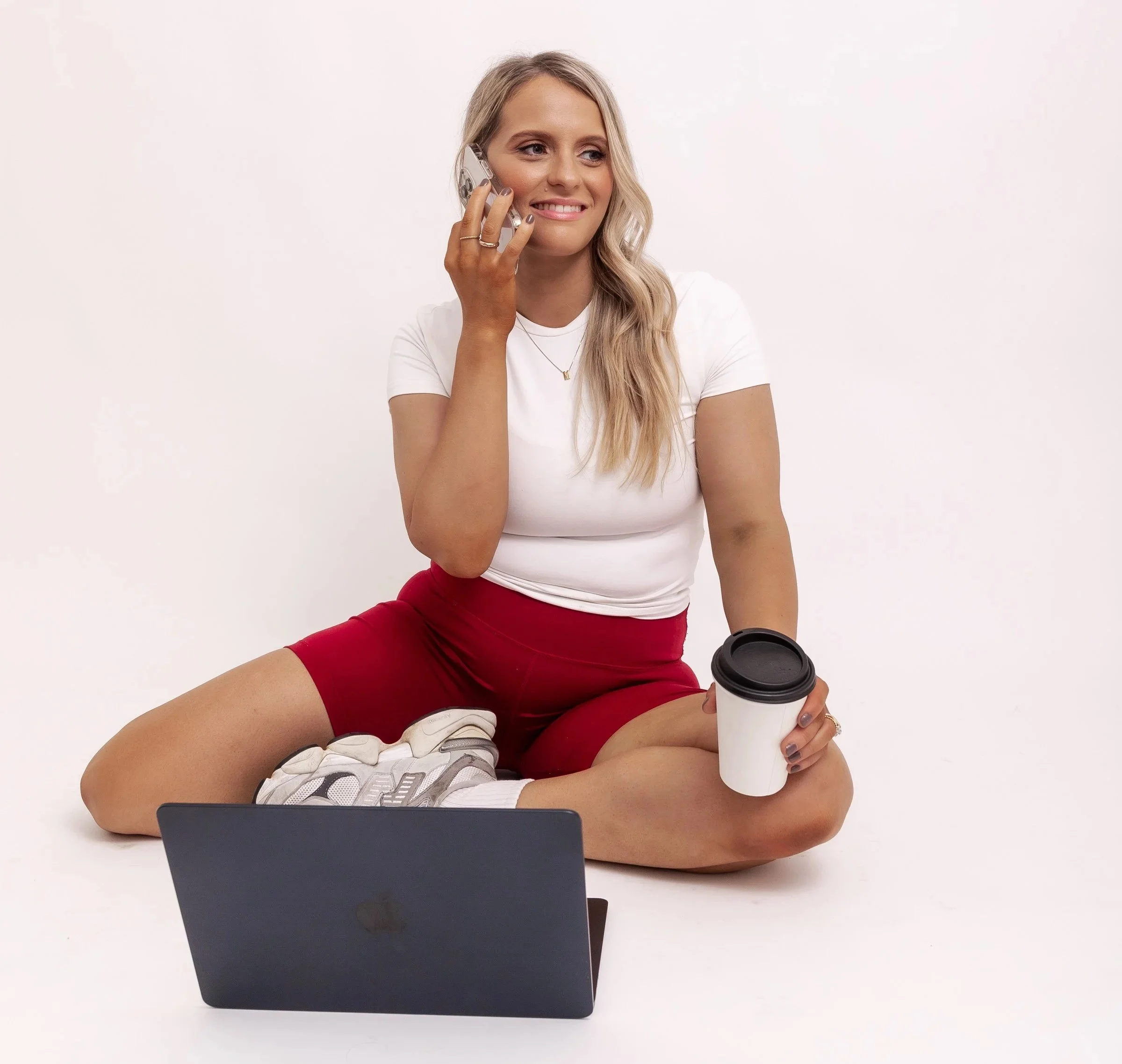A woman sitting cross-legged on the floor using a laptop, talking on her cellphone, holding a coffee cup, and dressed casually, against a plain white background.