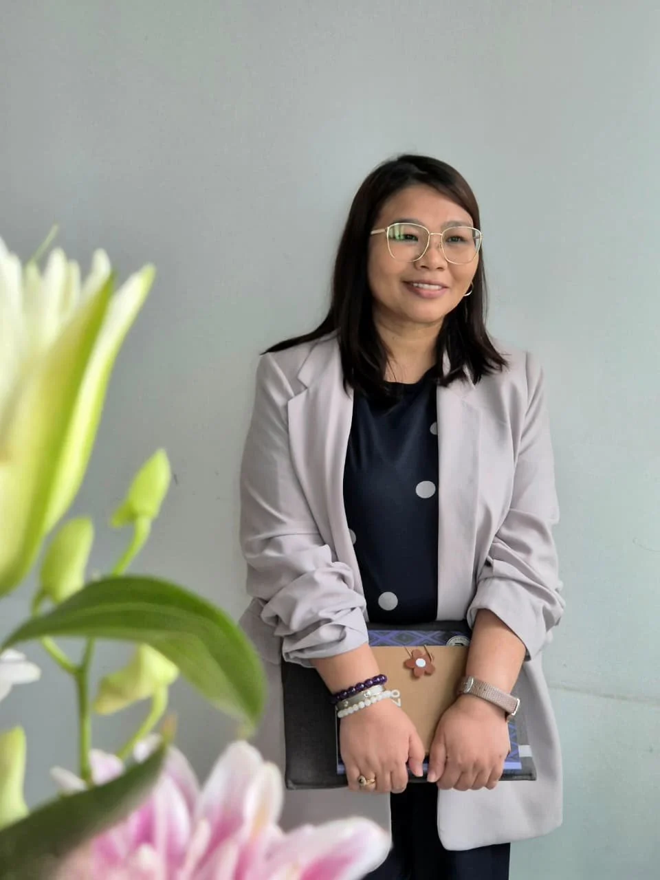 A woman with shoulder-length dark hair and glasses, wearing a light gray blazer over a black and white polka dot shirt, holding a notebook with a flower-shaped keychain, standing against a plain light-colored wall with colorful flowers visible in the foreground.