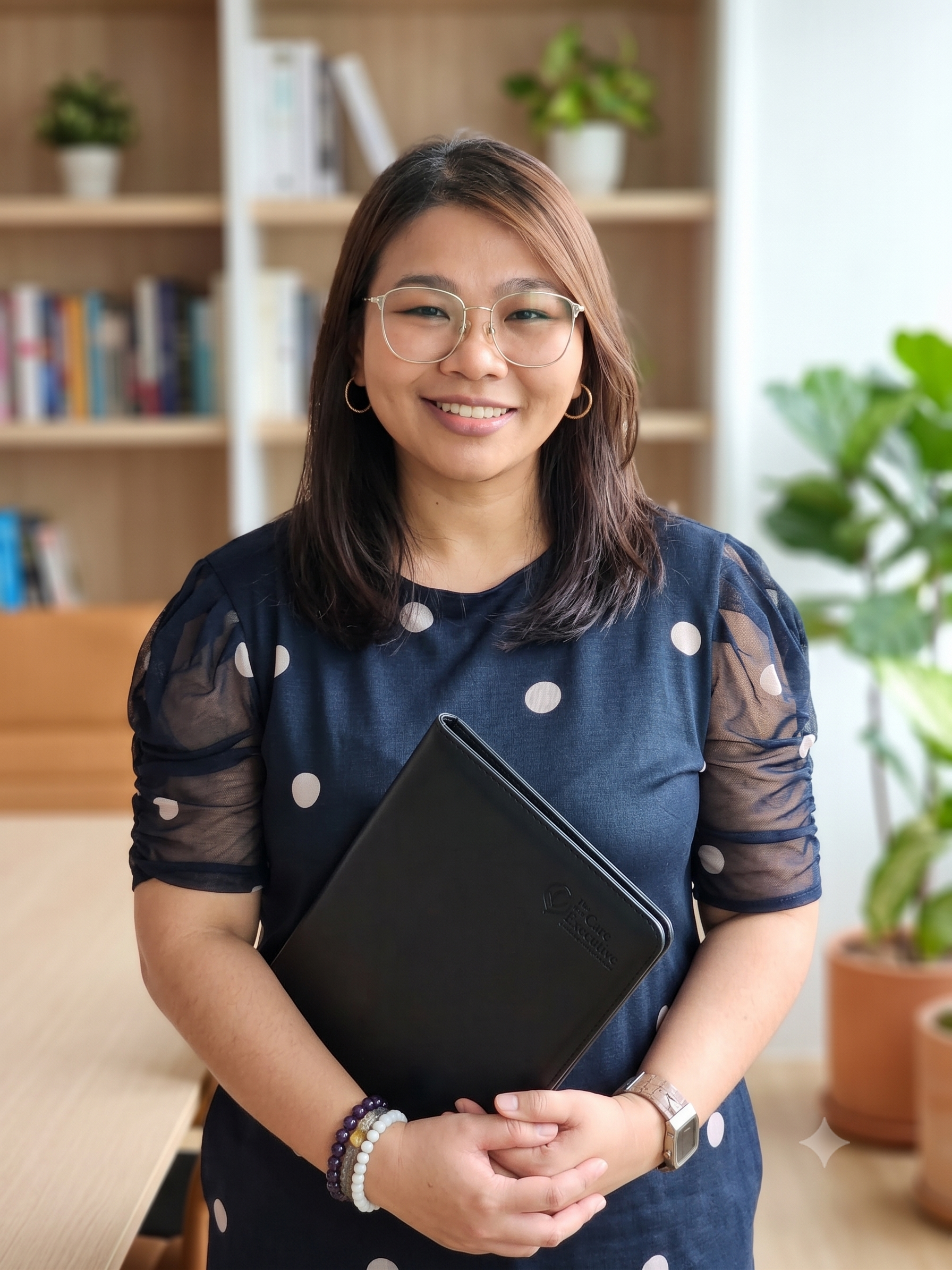 A woman with glasses and hoop earrings holding a black portfolio, wearing a navy blue dress with white polka dots, standing indoors with books and plants in the background.
