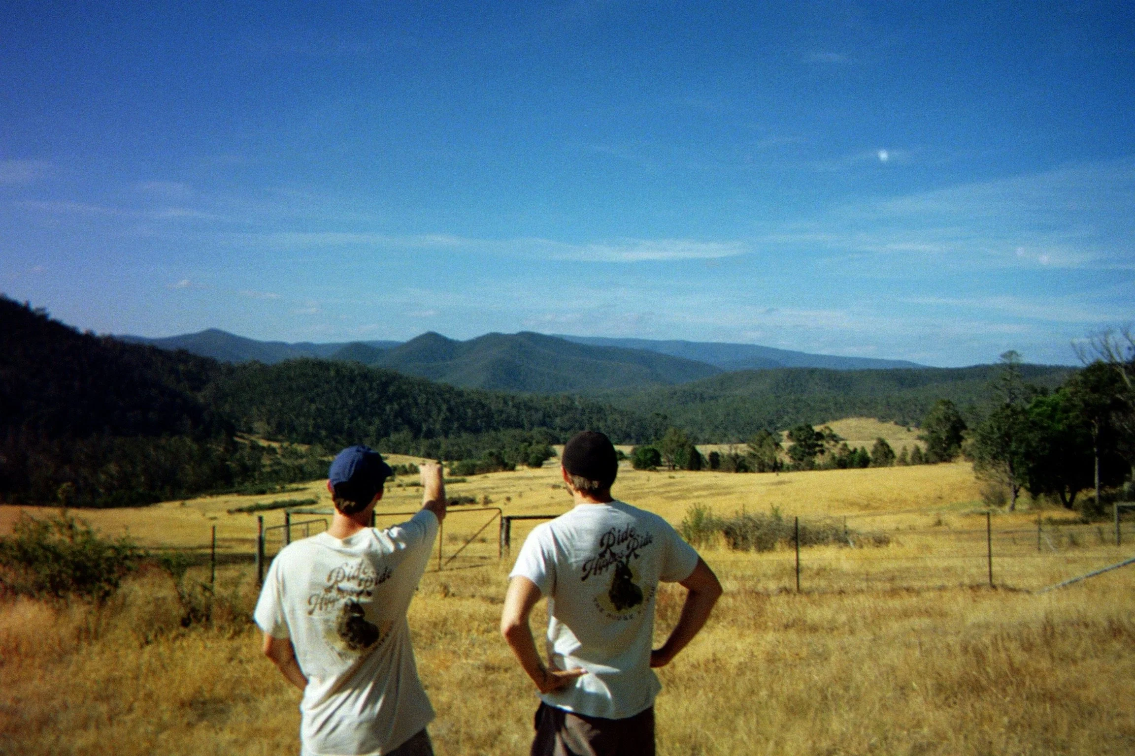 Two people standing outdoors in a field, with mountains and a blue sky in the background. One person is pointing towards the mountains.