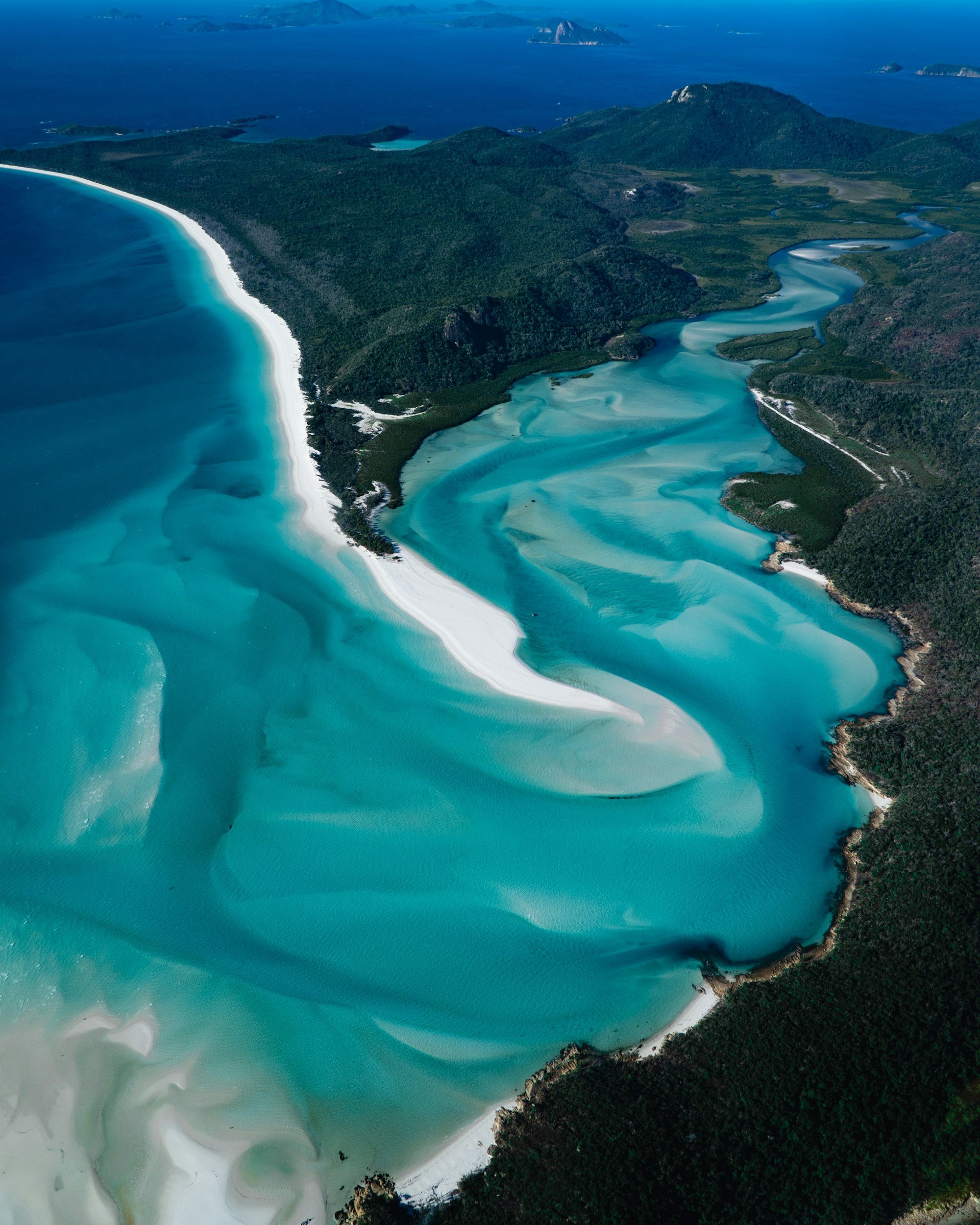 Aerial view of a turquoise bay with white sandy beaches, surrounded by lush green hills and small islands in the distance.