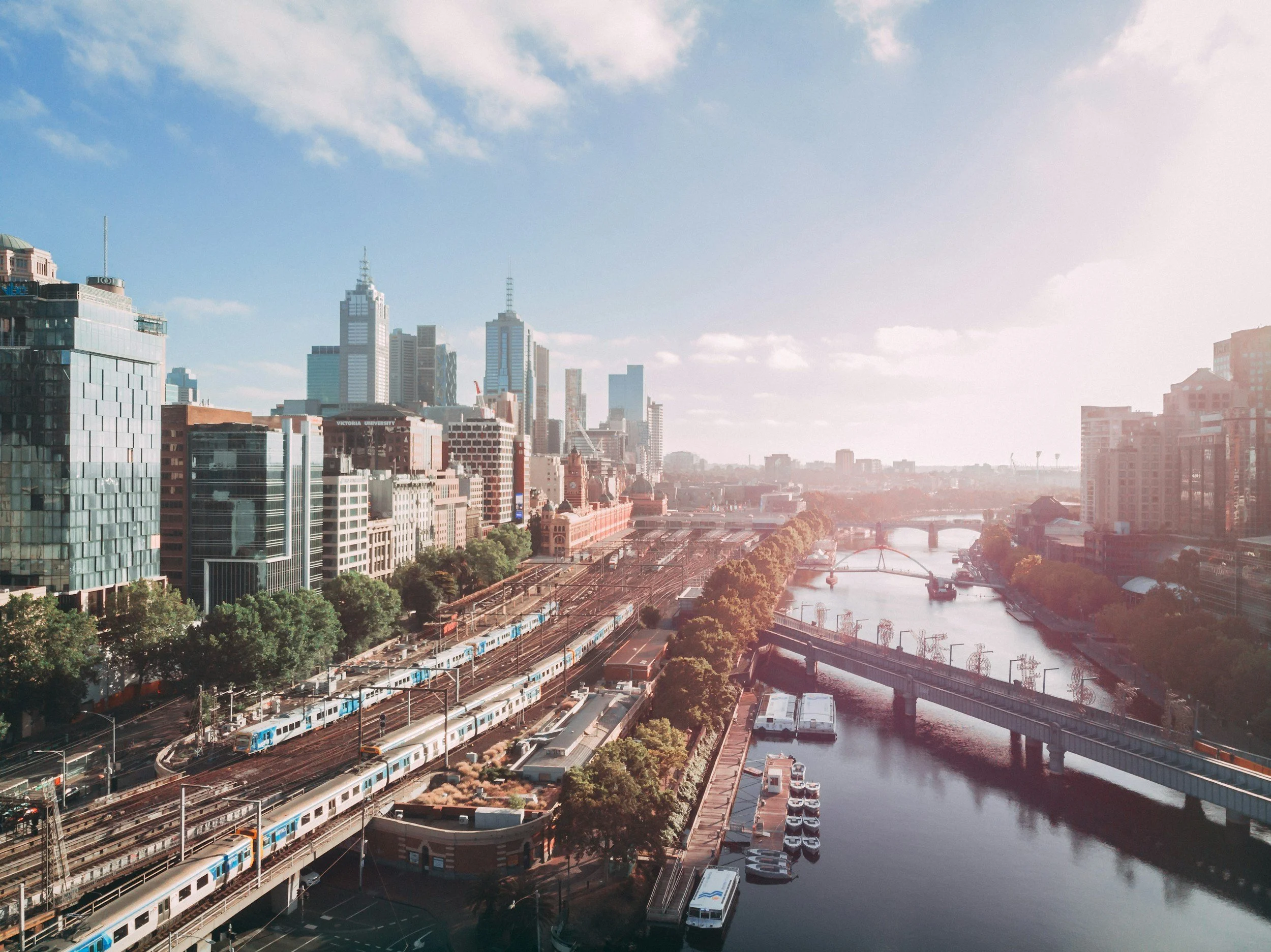 A cityscape featuring tall skyscrapers, a river with a bridge, railway tracks with trains, and a parking lot, under a partly cloudy blue sky.