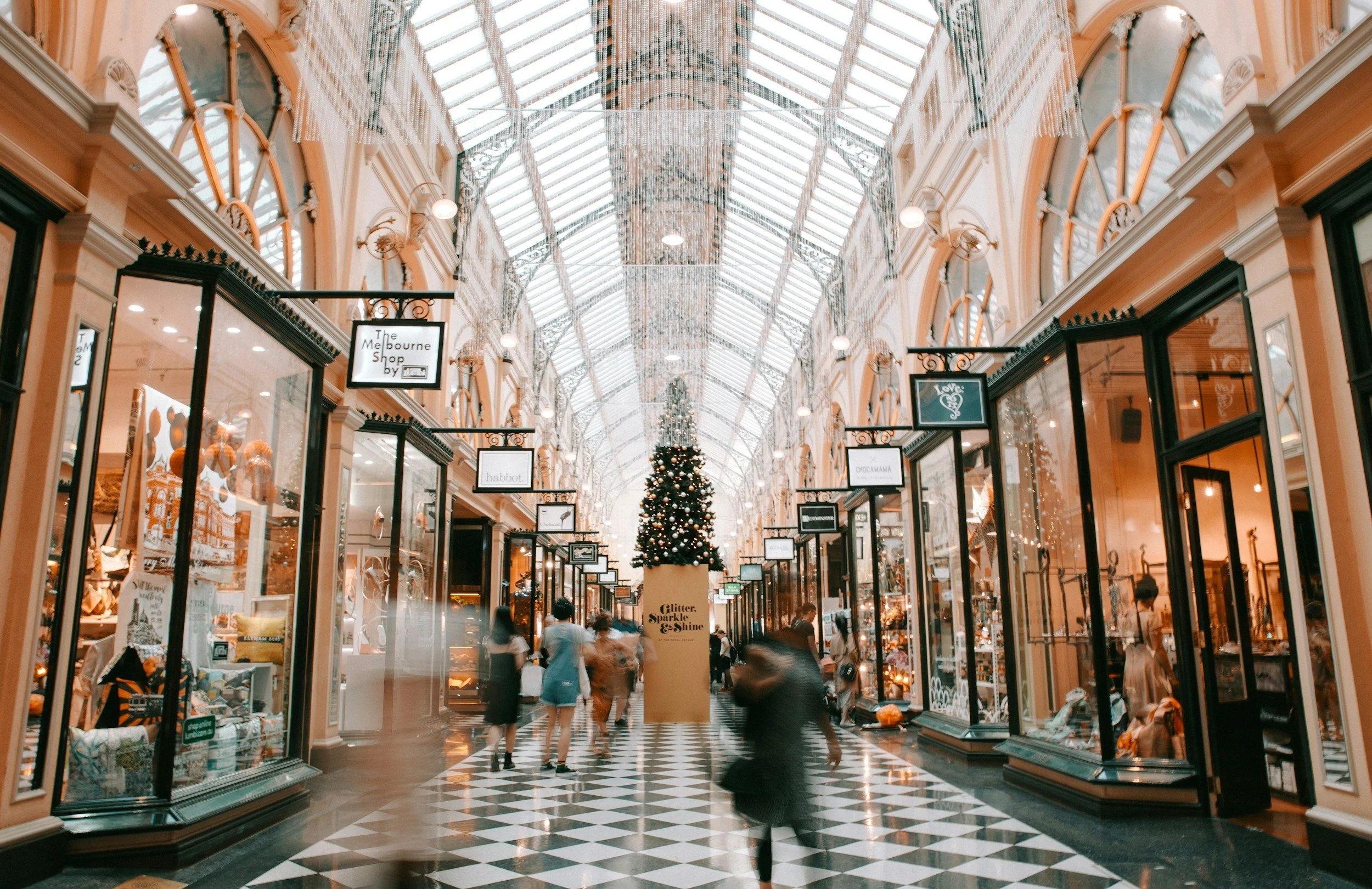 Interior of a shopping mall with a glass dome ceiling, storefronts, and a decorated Christmas tree in the center.