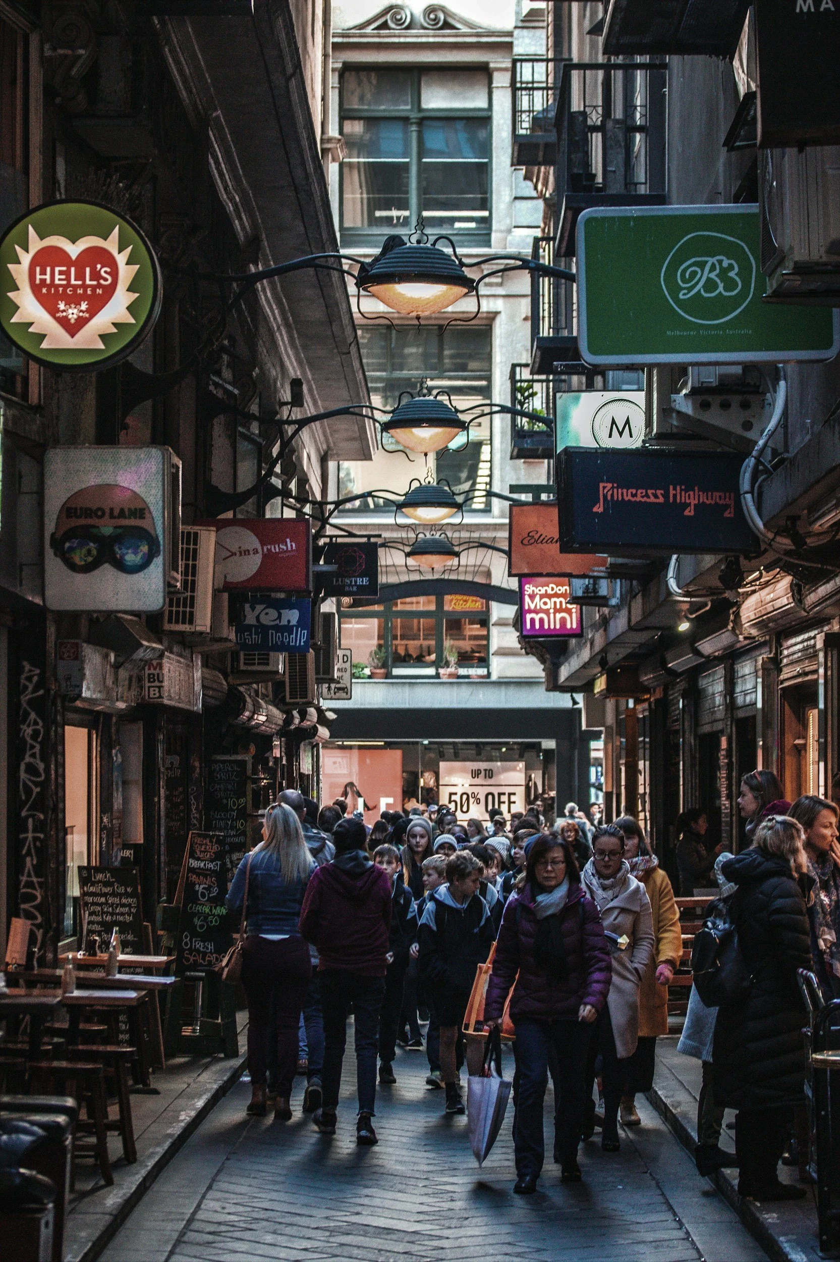A crowded city street with numerous signs, people walking, and outdoor cafes. The scene appears lively with various shops and a discount sign advertising up to 50% off.