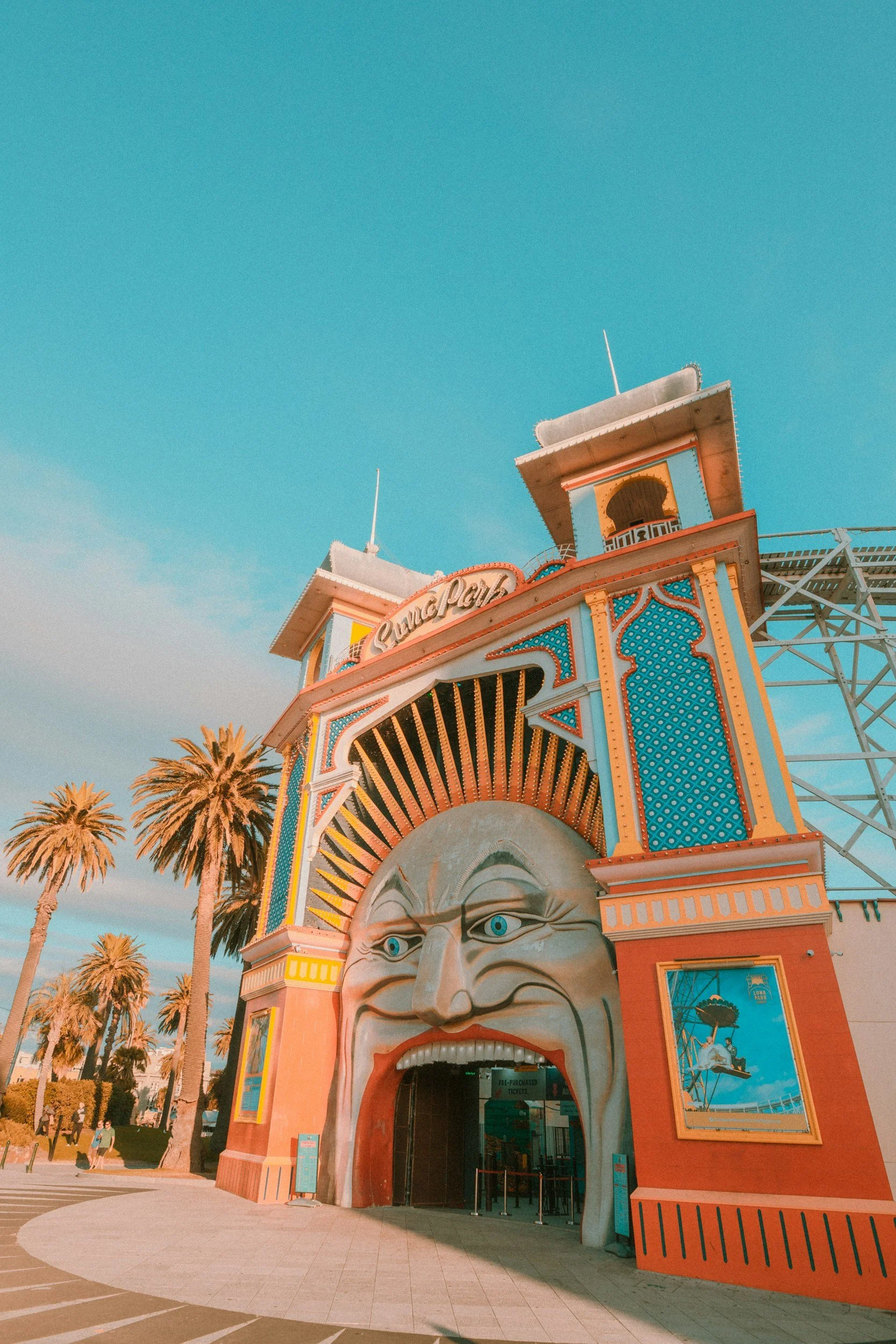 A colorful amusement park building with a large face sculpture on the front, featuring a pink mouth and blue eyes, with palm trees and a clear blue sky in the background.