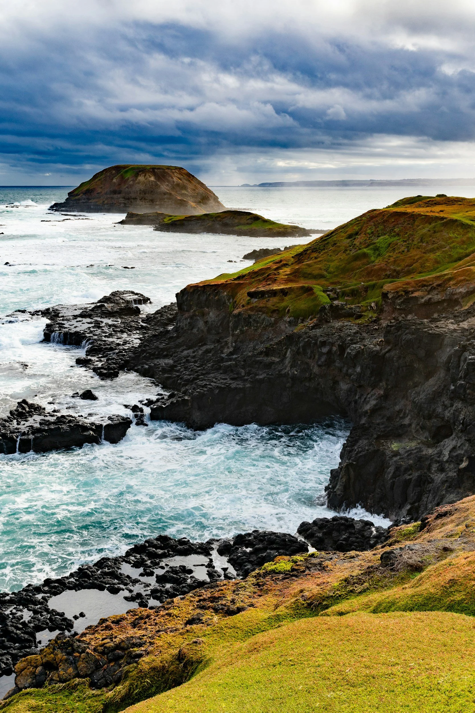 Coastal landscape with rocky shoreline, grassy hills, and a large rock formation in the ocean under a cloudy sky.