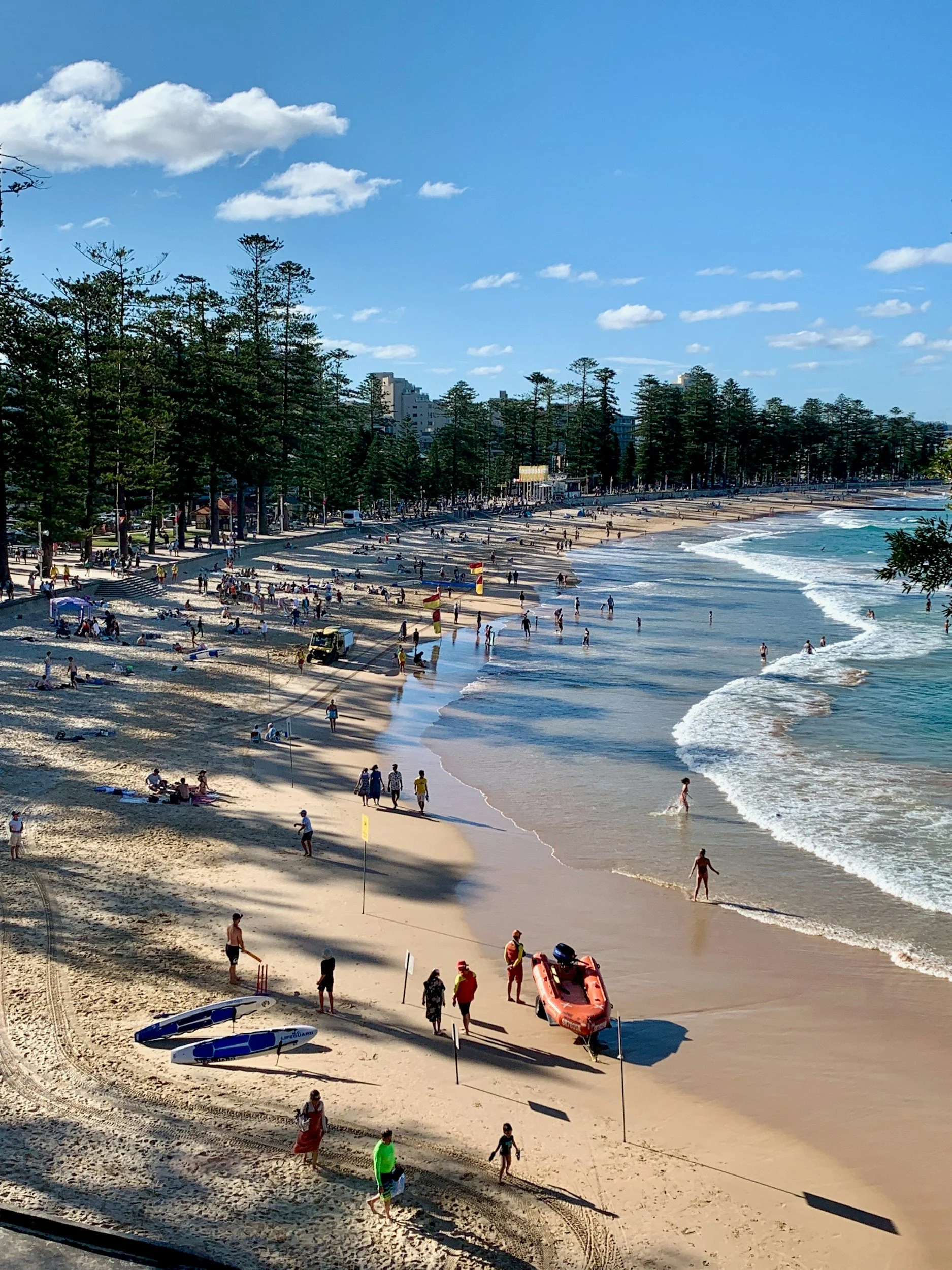 People enjoying a sunny day on the beach with umbrellas, lifeguard boats, and footprints in the sand, next to a row of trees and ocean waves.