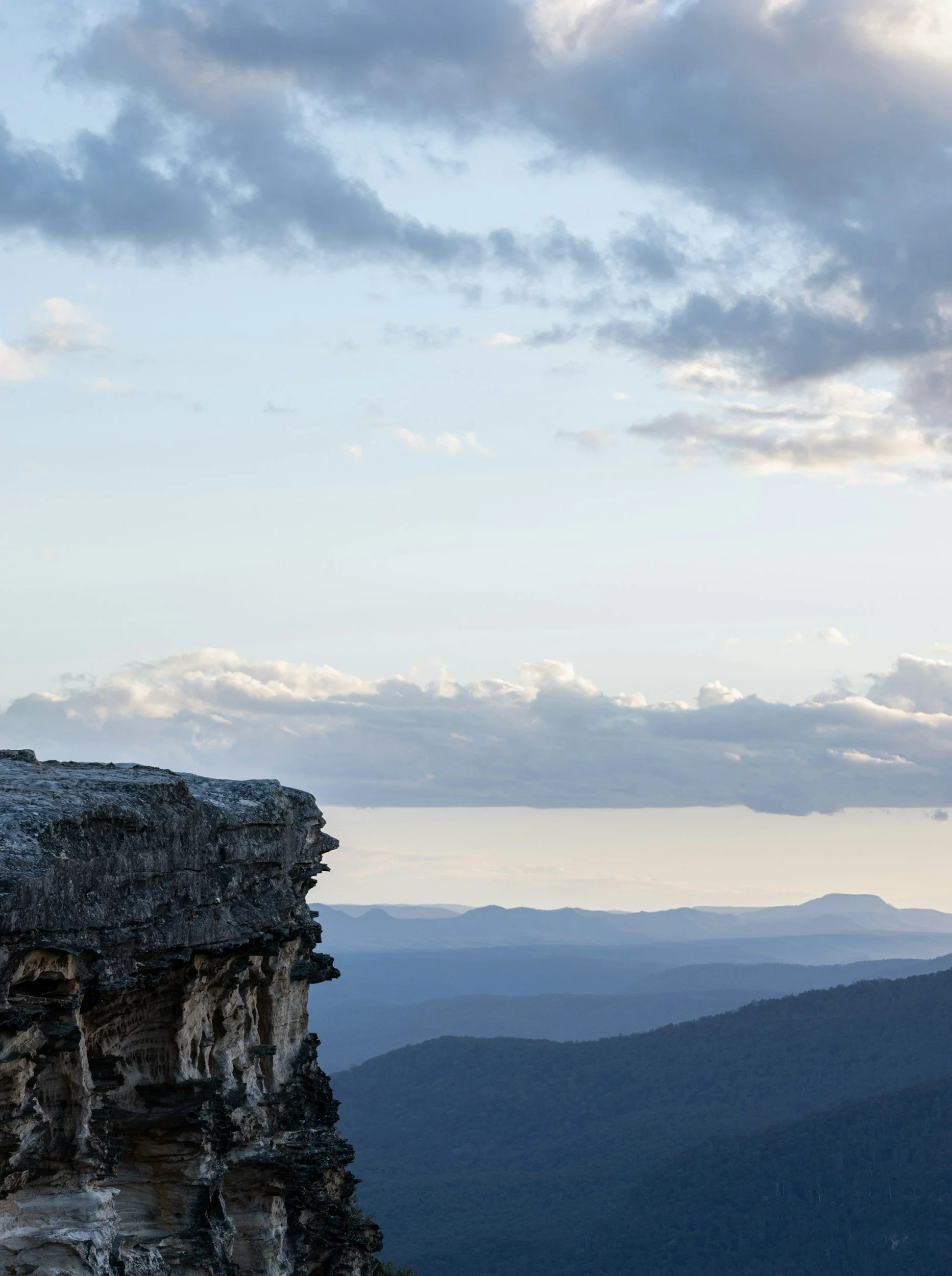 A rugged cliffside in the foreground with a distant mountain range and cloudy sky in the background.