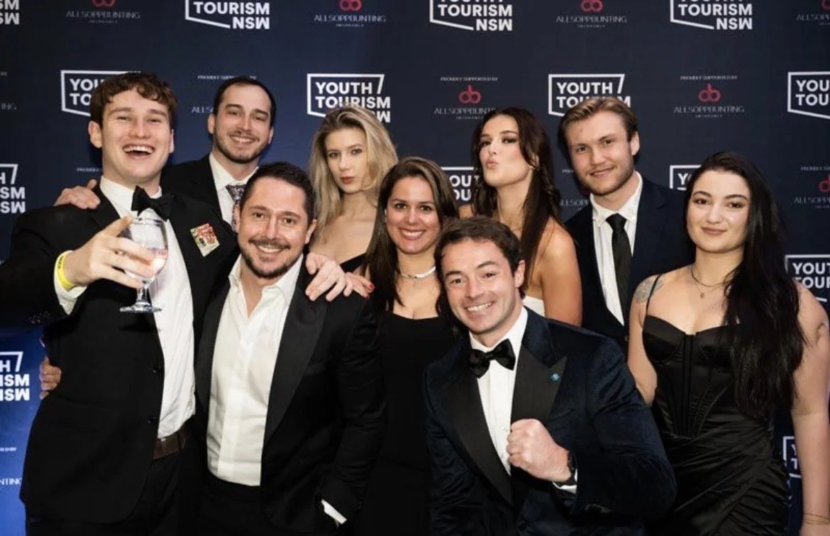 Group of young adults dressed in formal attire at a Youth Tourism NSW event, posing for a photo with a blue backdrop.