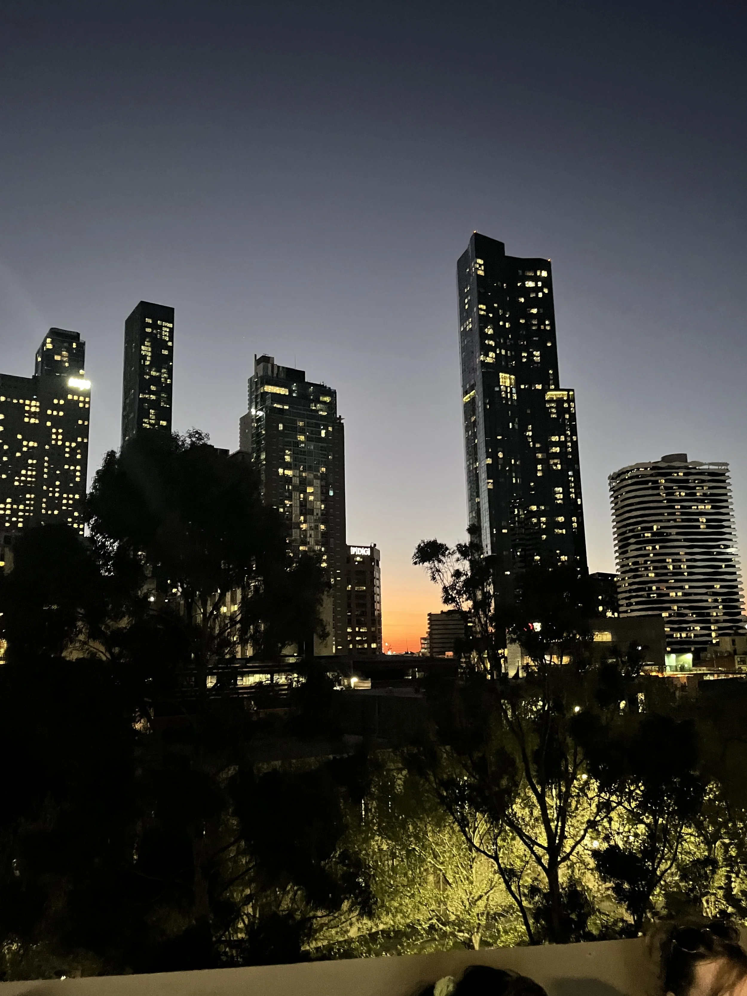 Nighttime cityscape with illuminated skyscrapers and a colorful sunset in the background.