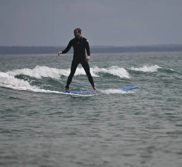 A man wearing a wetsuit standing on a stand-up paddleboard on the water, with small waves and a cloudy sky in the background.
