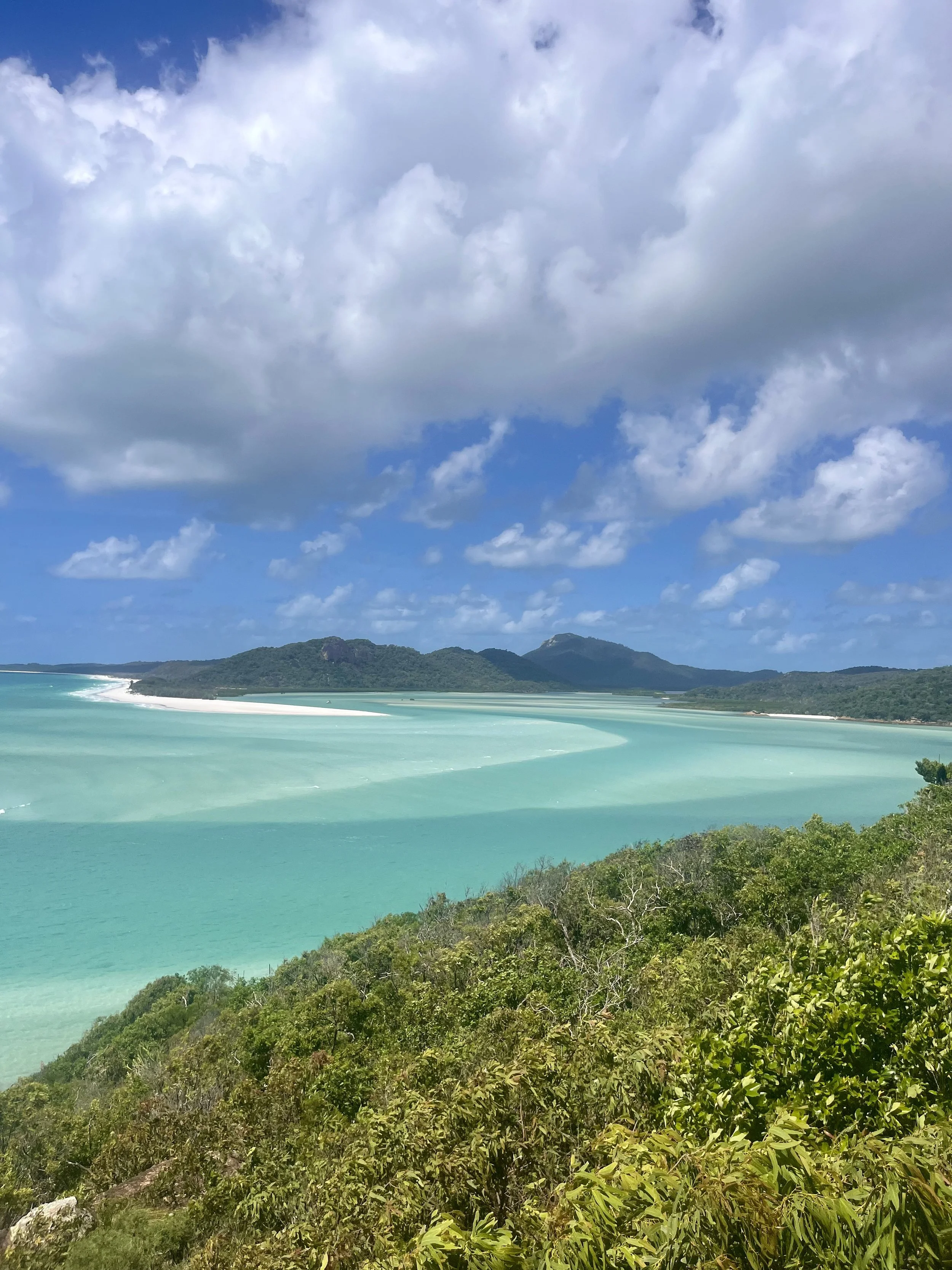 A scenic view of turquoise ocean water meeting a white sandy beach at the base of green hills under a partly cloudy sky.