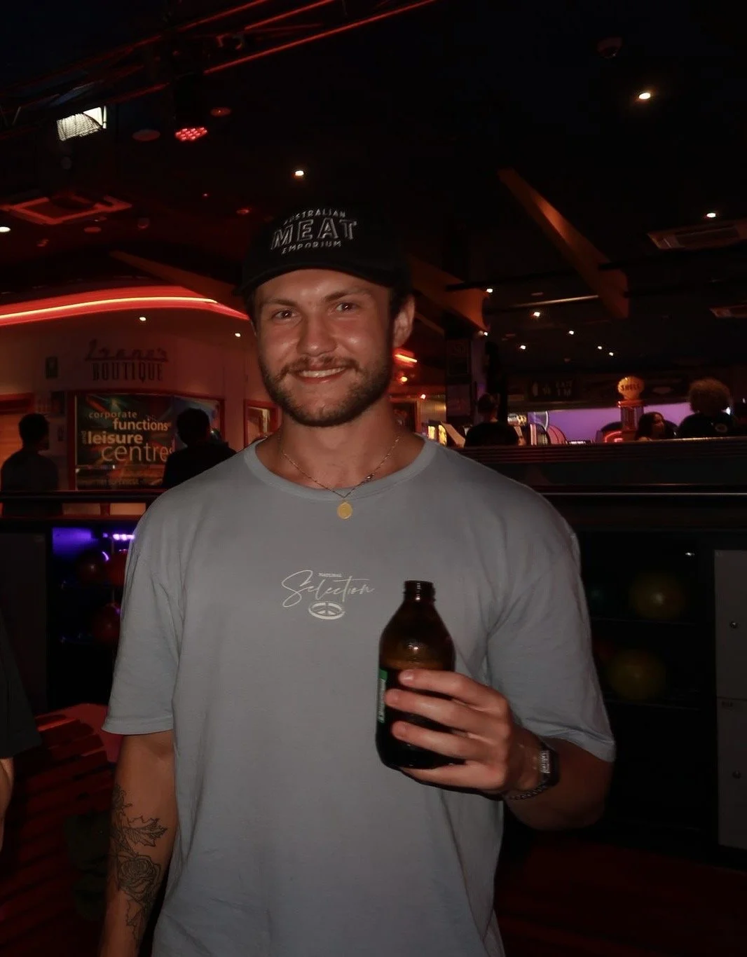 A smiling man holding a brown bottle in a dimly lit bar or casino with colorful lights and patrons in the background.