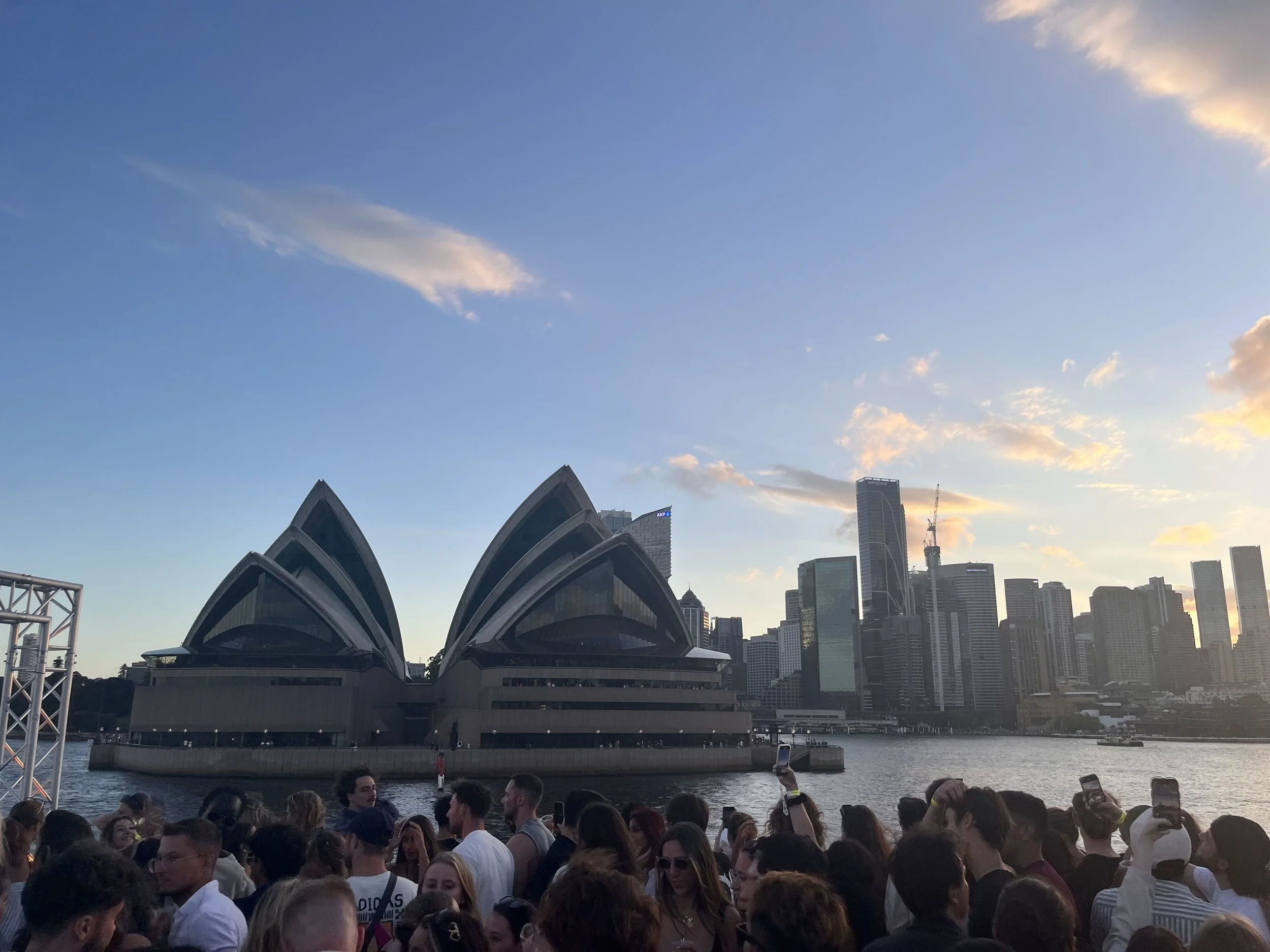 A large crowd of people gathered near a body of water with the Sydney Opera House and city skyline in the background during sunset.