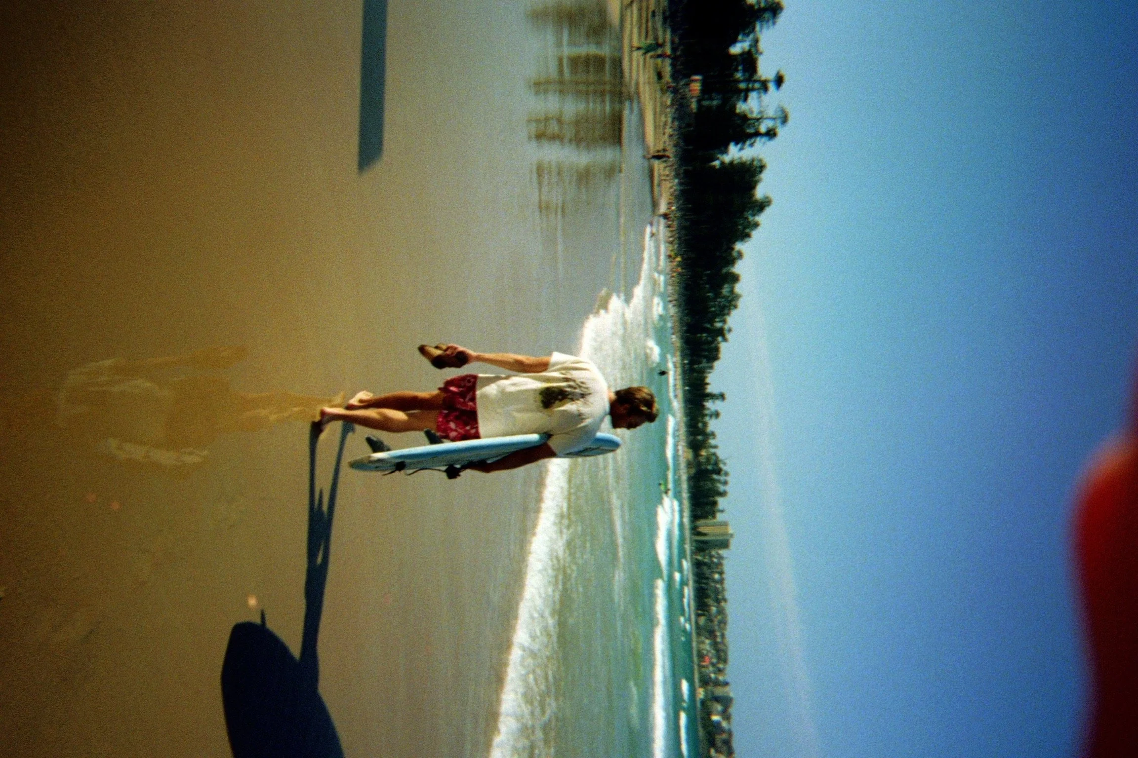 Person in a white shirt and red shorts walking on a beach while carrying a surfboard, with ocean waves and distant trees in the background.