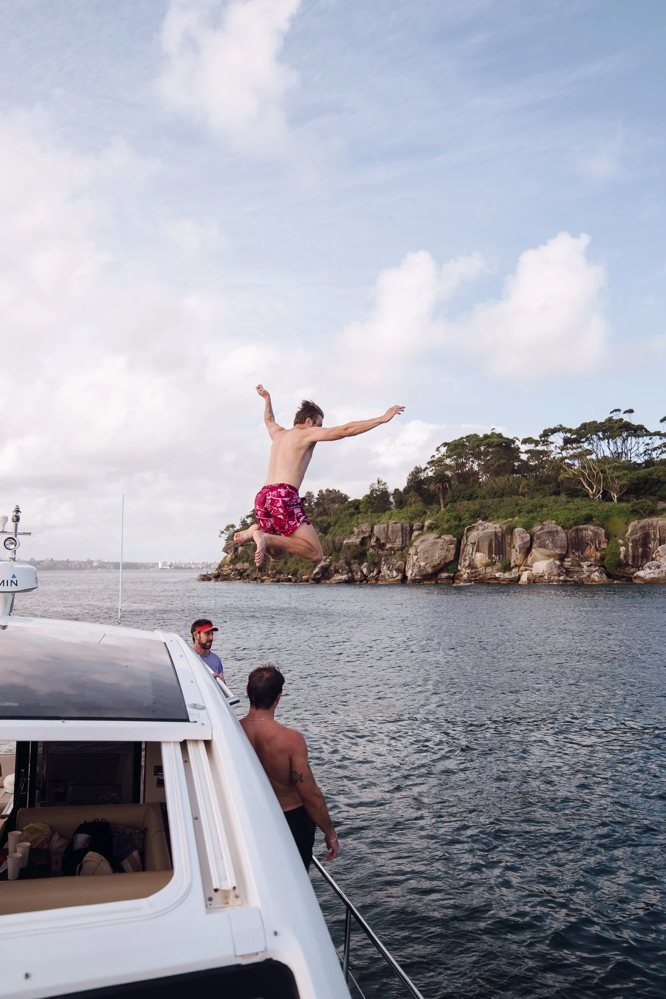A young man in red shorts is mid-air jumping off a boat into the water near a rocky shoreline with trees. Two other men are on the boat, one leaning over the side and the other standing behind him, looking at the jumper. The sky is partly cloudy, and the water appears calm.