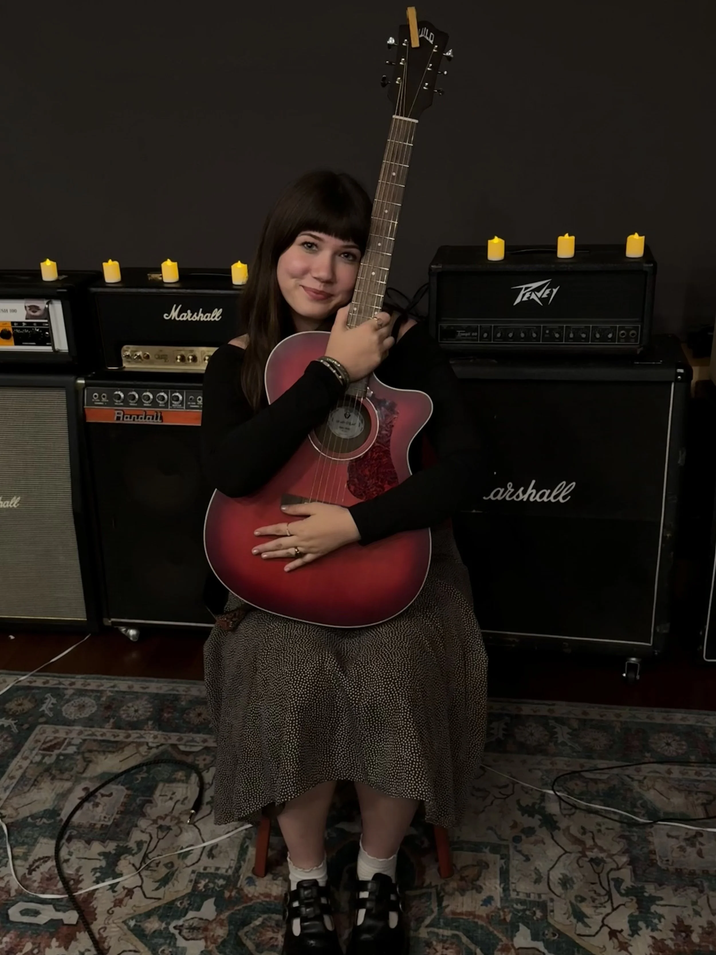 A woman sitting in front of guitar amplifiers, holding an acoustic guitar with a pink and red color gradient, wearing a black long sleeve top and a patterned skirt, socks, and black sandals, with candles on the amplifiers and a patterned rug on the floor.