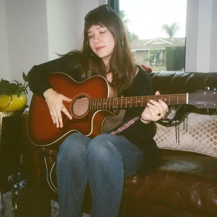 Young woman with long brown hair playing an acoustic guitar indoors near a window.