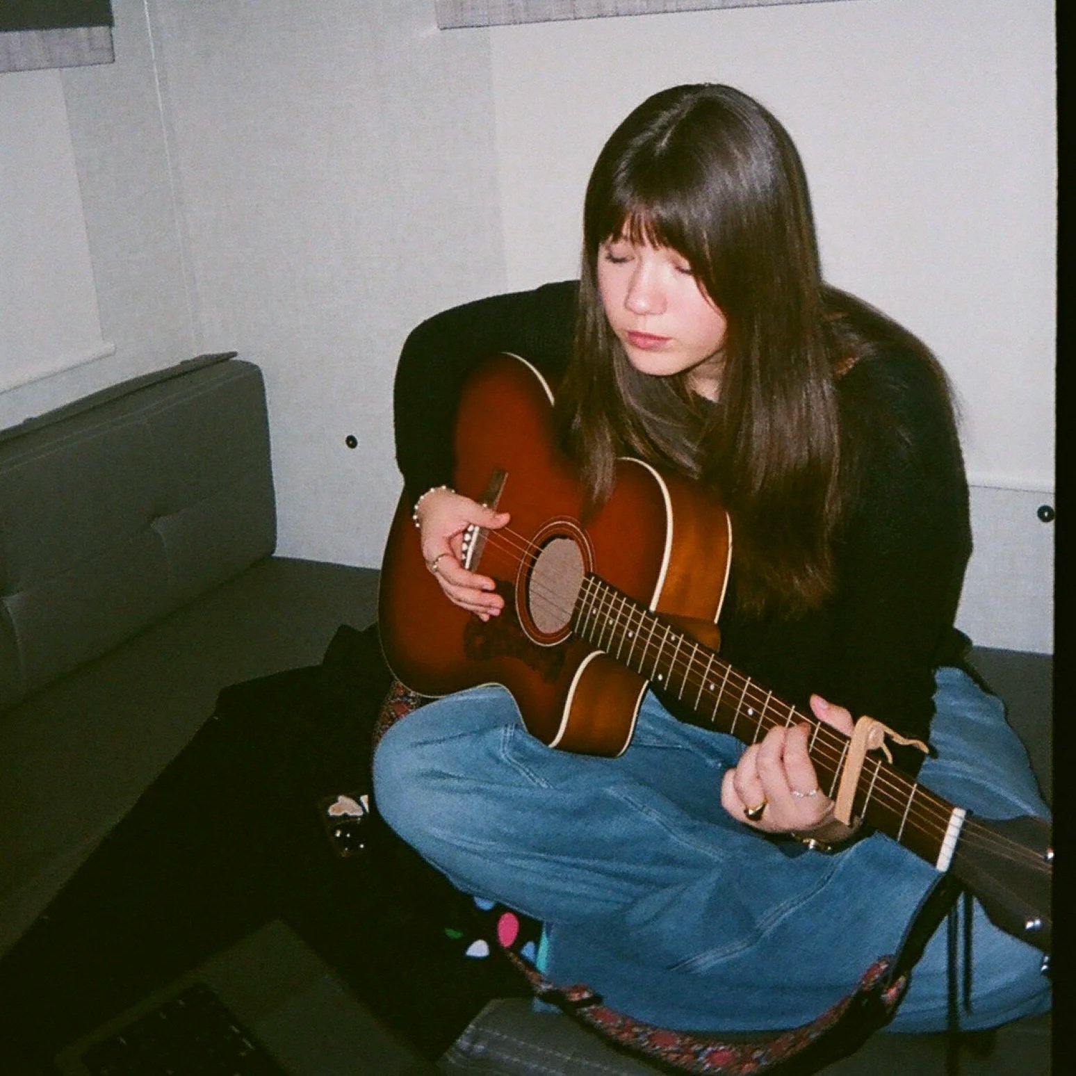 A young woman with long dark hair sitting on a patterned chair, wearing a black sweater and blue jeans, playing an acoustic guitar in a room with white walls and black flooring.