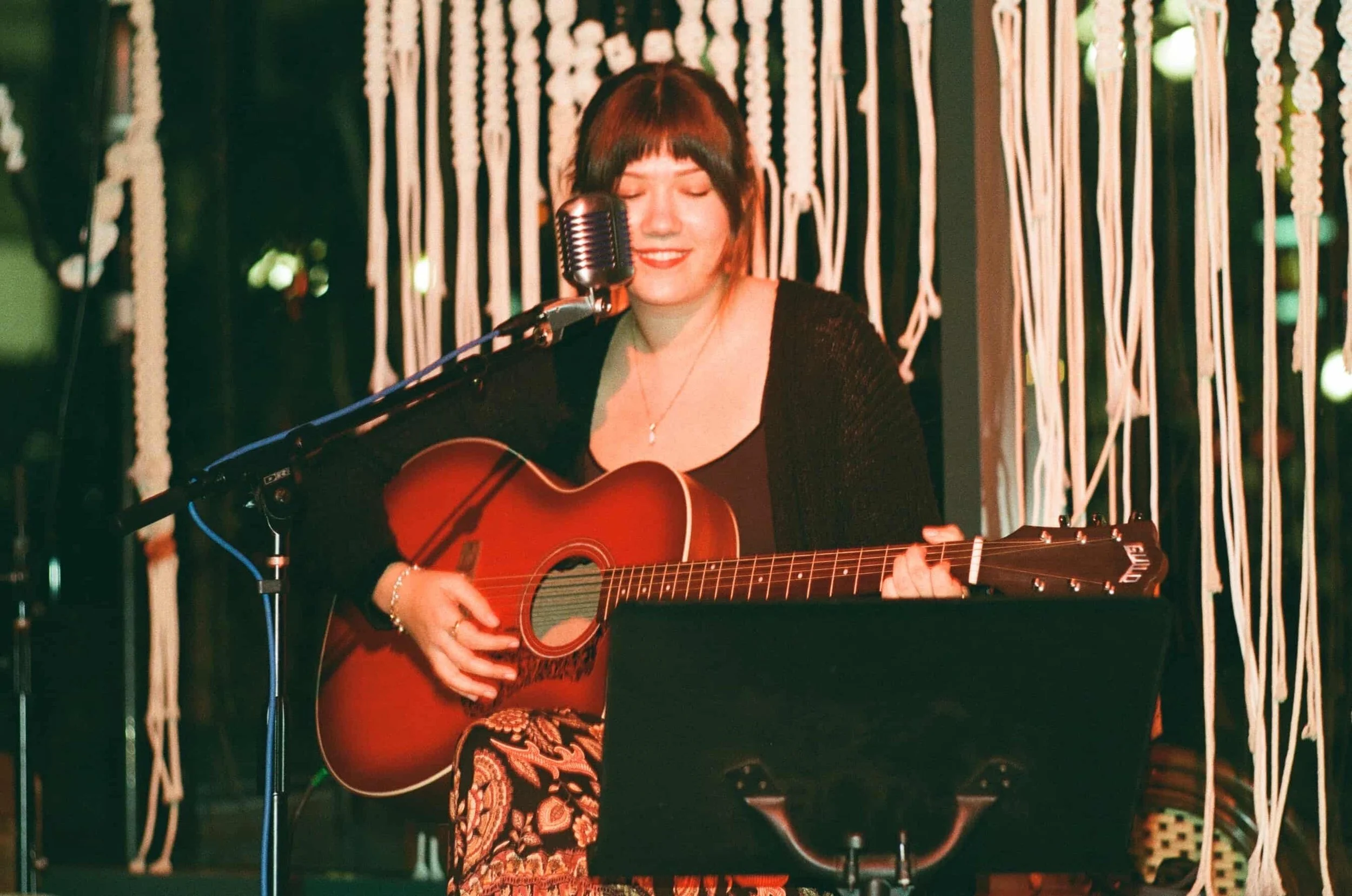 A woman with short hair and red lipstick is playing an acoustic guitar and singing into a vintage microphone. She has her eyes closed and appears to be enjoying the music. She is in a cozy, decorated indoor space with a background of hanging rope or string decorations.