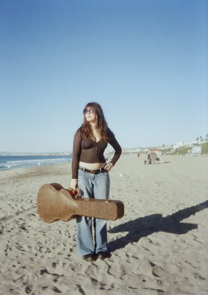 Woman standing on the beach wearing sunglasses, black long-sleeve top, blue jeans, holding a guitar case in her right hand, with the ocean and sandy beach in the background.