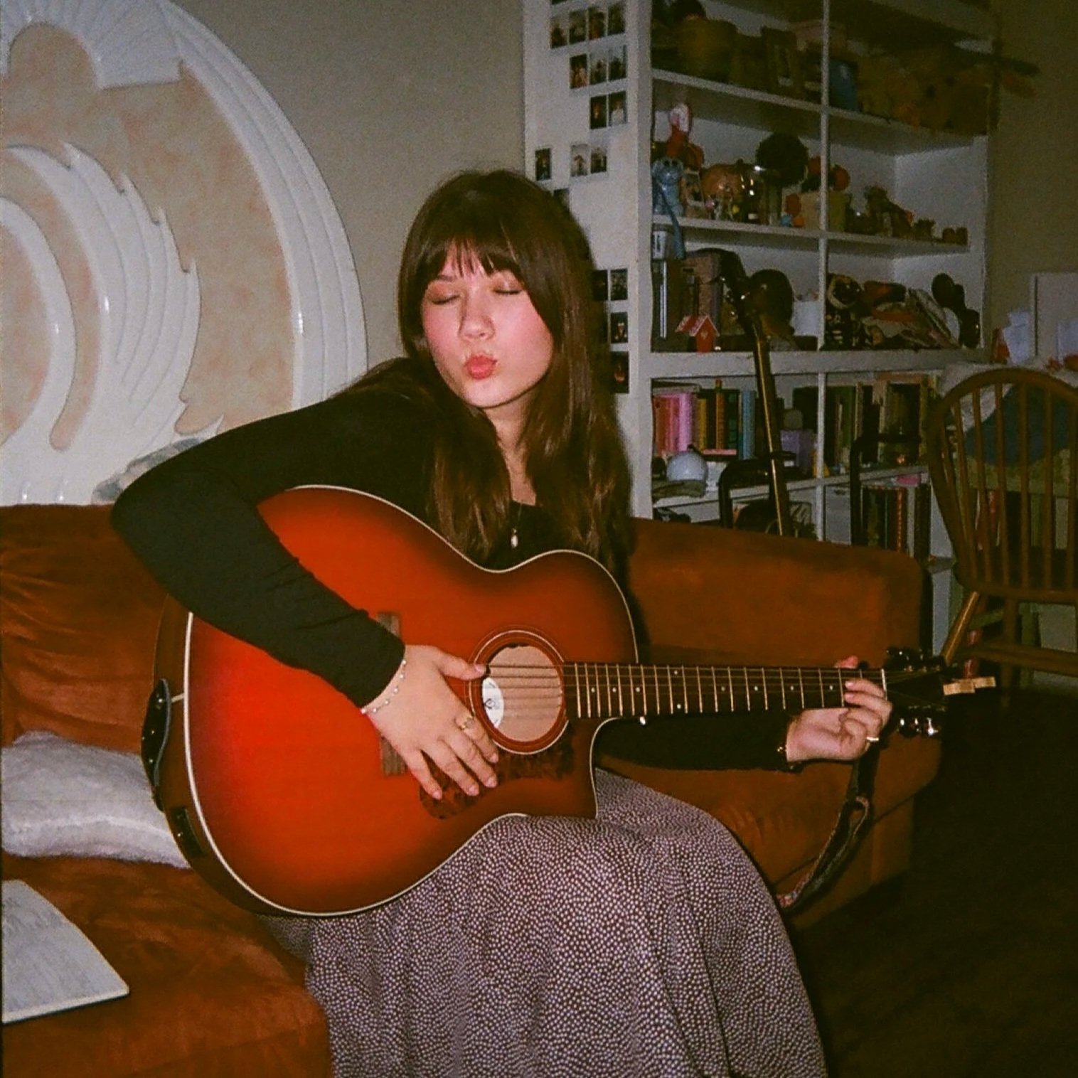 A woman with long brown hair and bangs is sitting on a brown couch, playing an acoustic guitar. She is wearing a black long-sleeve top and patterned pants, and her eyes are closed as she sings or makes a gesture with her lips. Behind her is a white bookshelf filled with books and decorative items, and to the right of the bookshelf is a wooden chair. The room has beige walls and a decorative white wall molding.