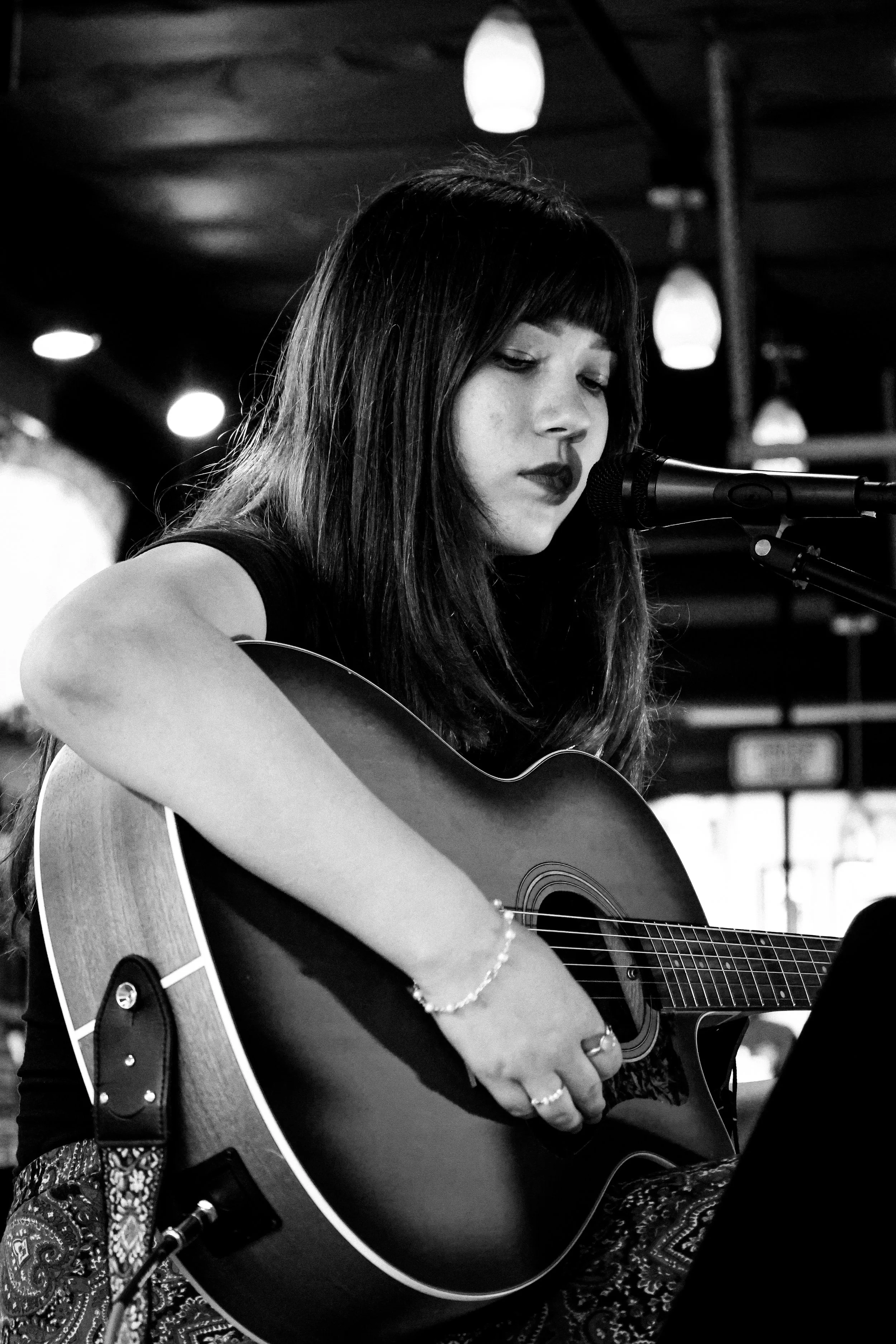 A woman with dark hair and bangs playing an acoustic guitar and singing into a microphone at what appears to be a live music venue.