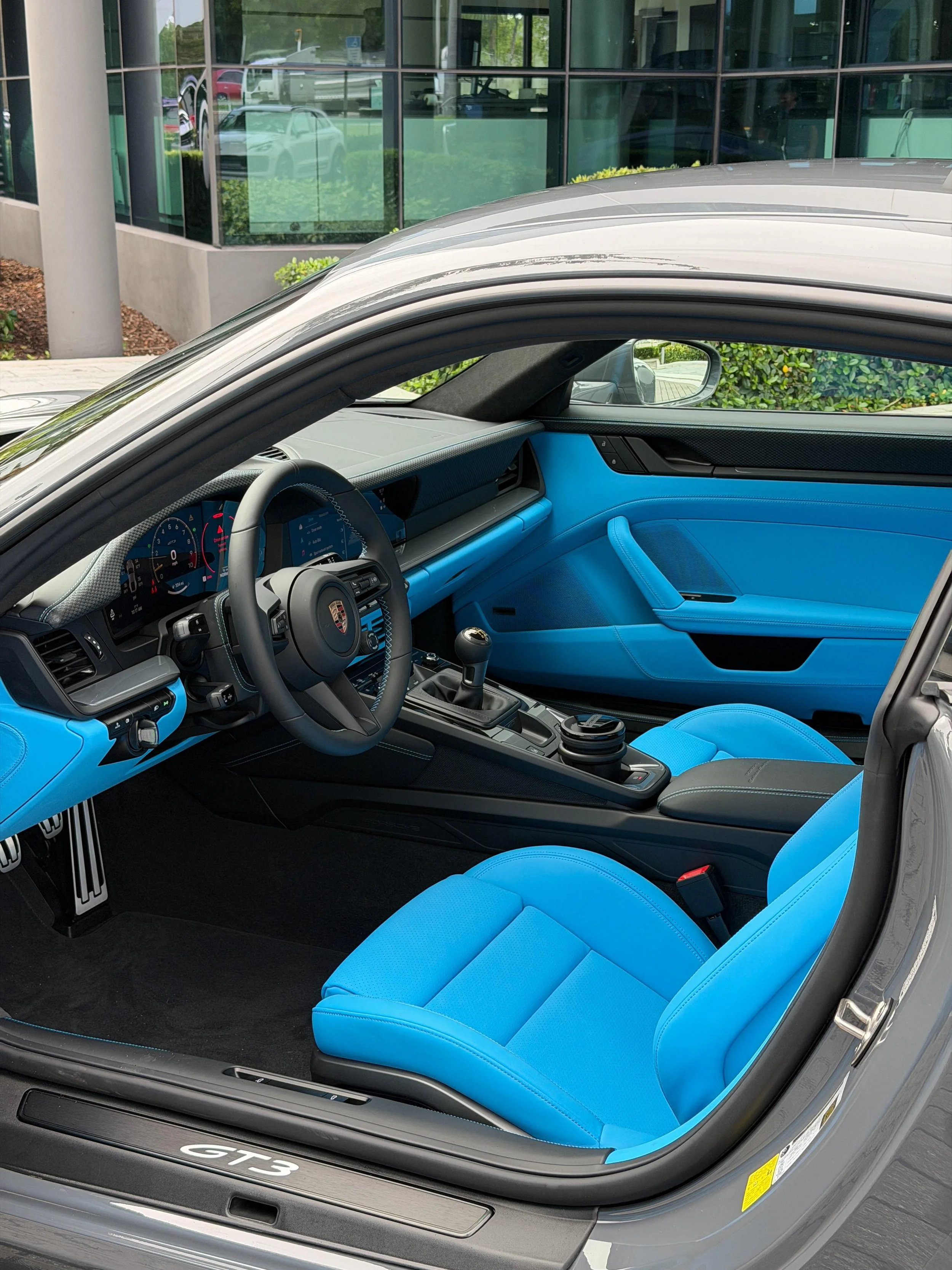 The image shows the interior of a gray Porsche GT3 sports car with bright blue seats, parked outside a building with large glass windows, reflecting trees and other cars.