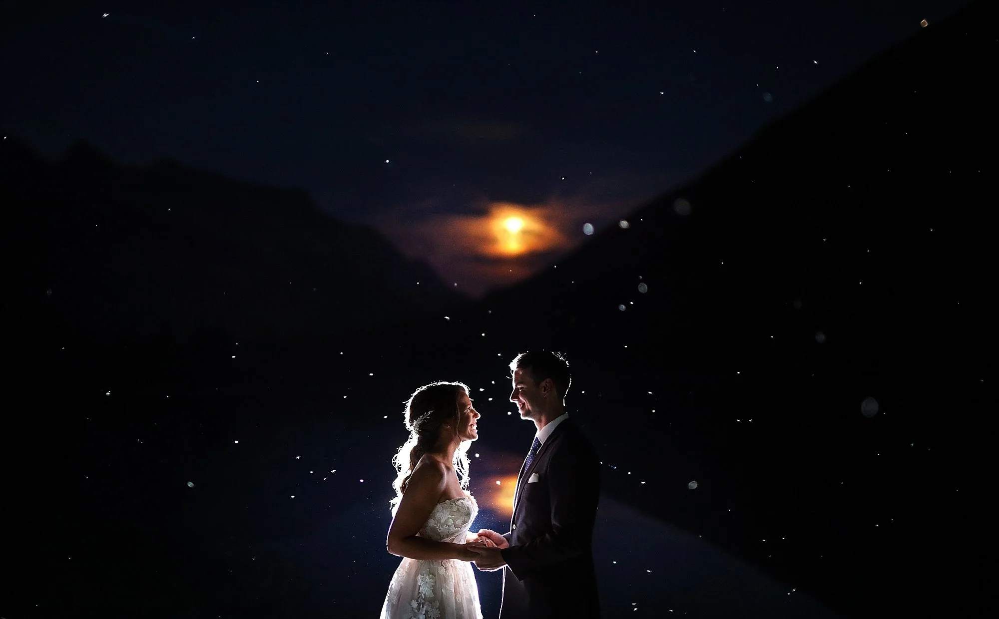 A couple dressed in wedding attire holding hands and smiling at each other outdoors at night, with a dark sky, stars, a bright moon, and mountains in the background.