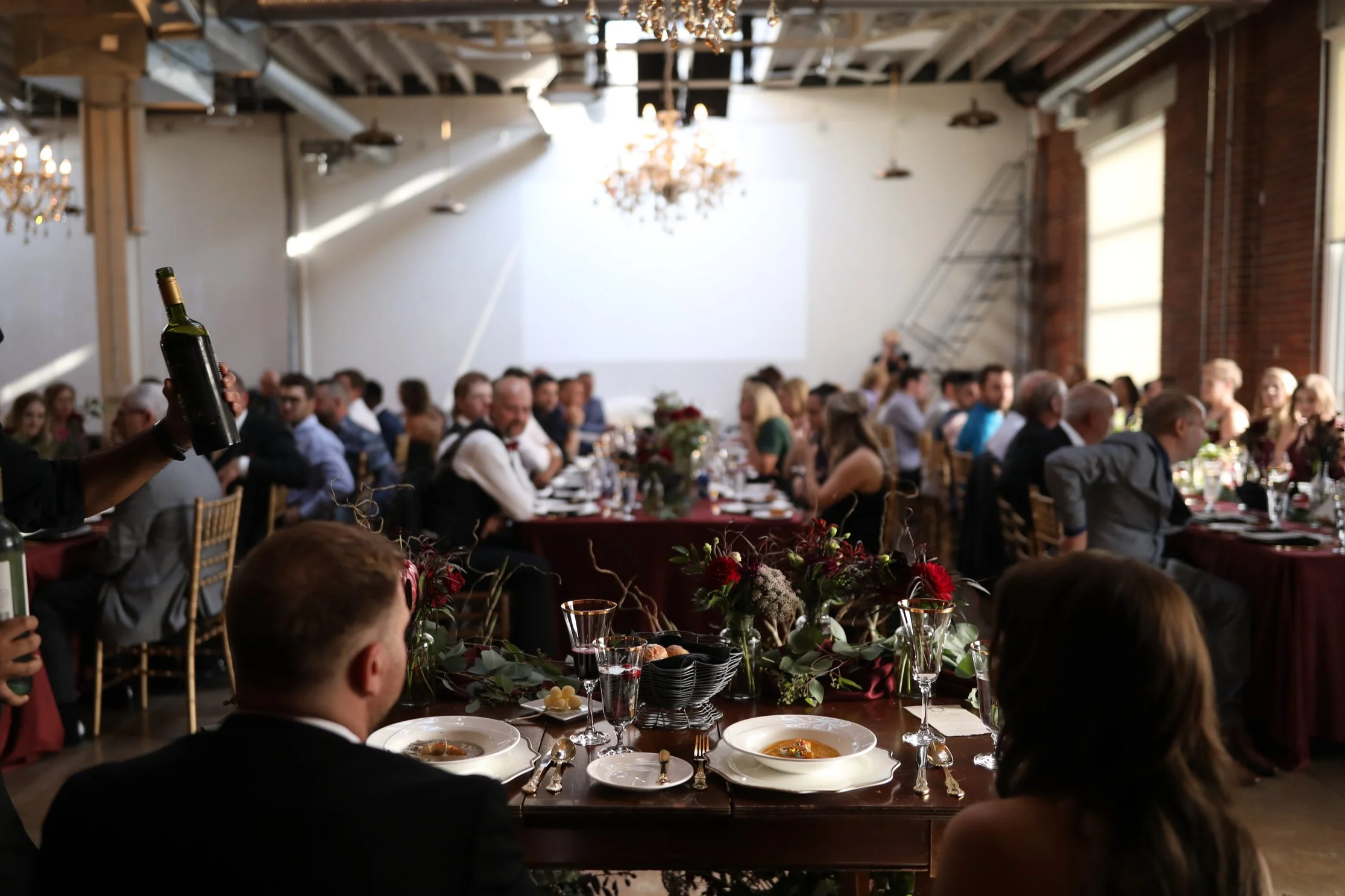 A large gathering of people seated at long tables decorated with floral arrangements, dining at a banquet-style event in an industrial-style venue with exposed brick walls, chandeliers, and a large blank screen on the wall.