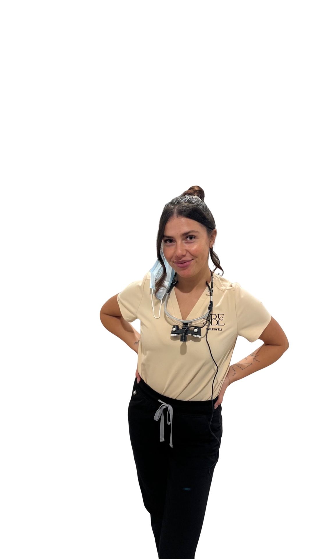 A young woman with dark hair in a bun, wearing a beige medical scrub top and black pants, stands with her hands on her hips, smiling at the camera. She has a headlamp, a face mask hanging around her neck, and some tattoos on her arms.