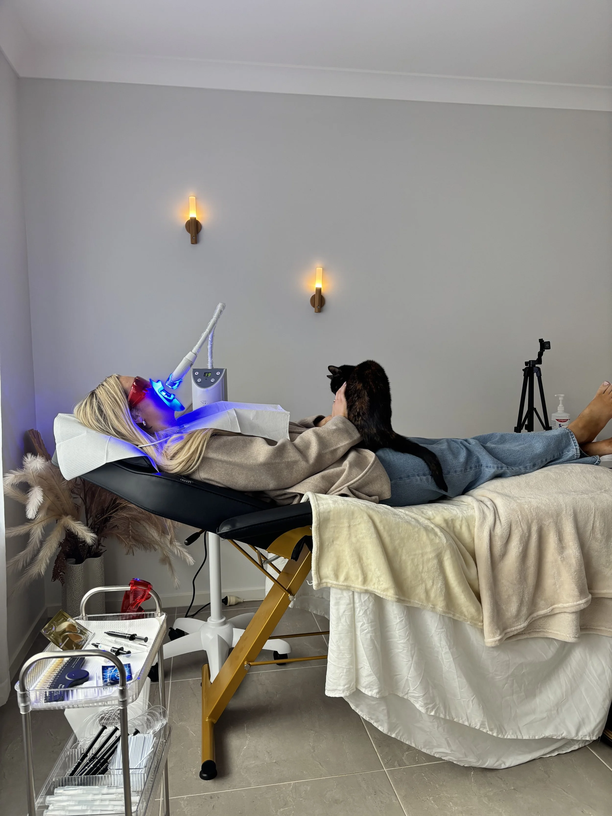 A woman is lying on a medical examination table with a black cat on her chest, in a room with light-colored walls and decorative wall lights. Medical equipment, including a laser device, is nearby on a small cart and the floor has various medical instruments.