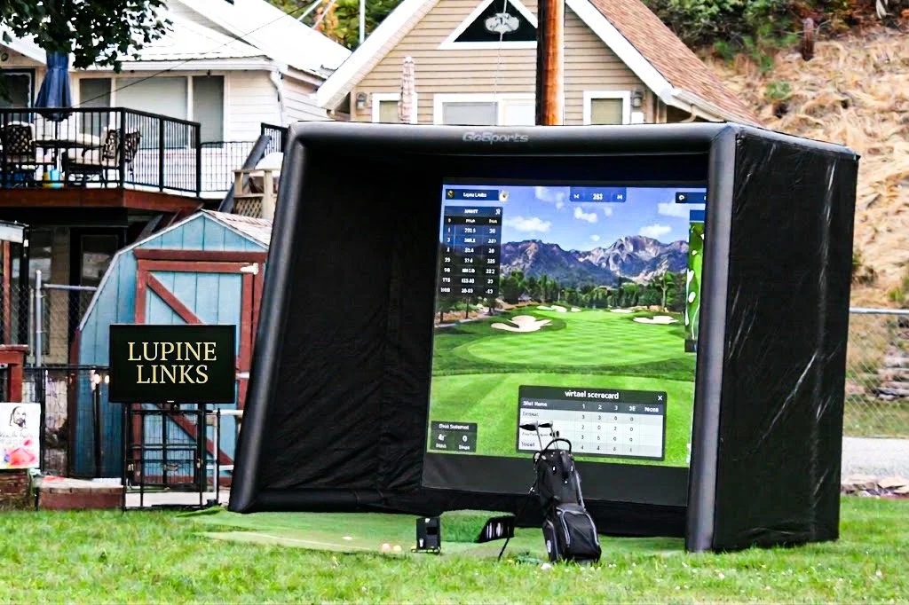 A golf simulator setup outdoors on a grassy area, with a large screen displaying a golf course landscape. There is a golf bag and club on the ground in front of the screen. In the background, there are houses and a hillside.