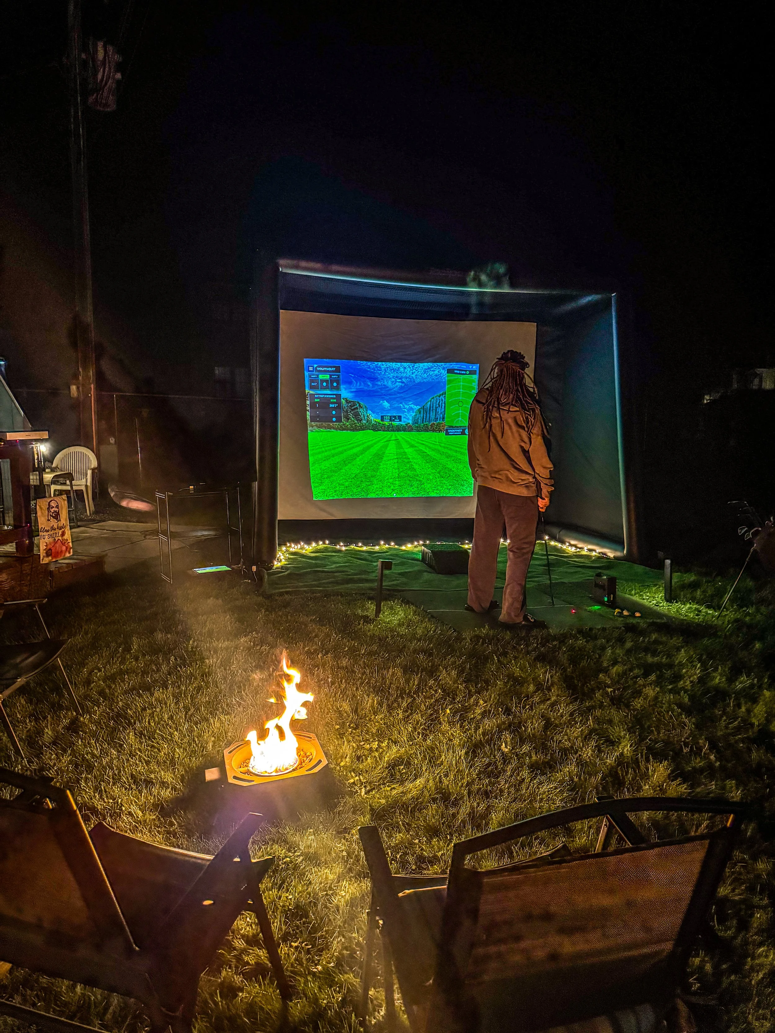 Person standing in front of an outdoor movie screen displaying a soccer game at night, surrounded by chairs, a small fire, and outdoor lighting.