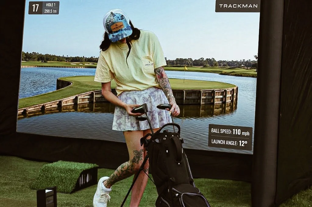 A woman standing in front of a golf simulator screen, holding a golf club and looking at the screen showing a virtual golf course with water hazards. She is wearing a yellow t-shirt, a patterned skirt, a cap, and has tattoos on her legs and arms. A golf bag with clubs is on the ground in front of her.