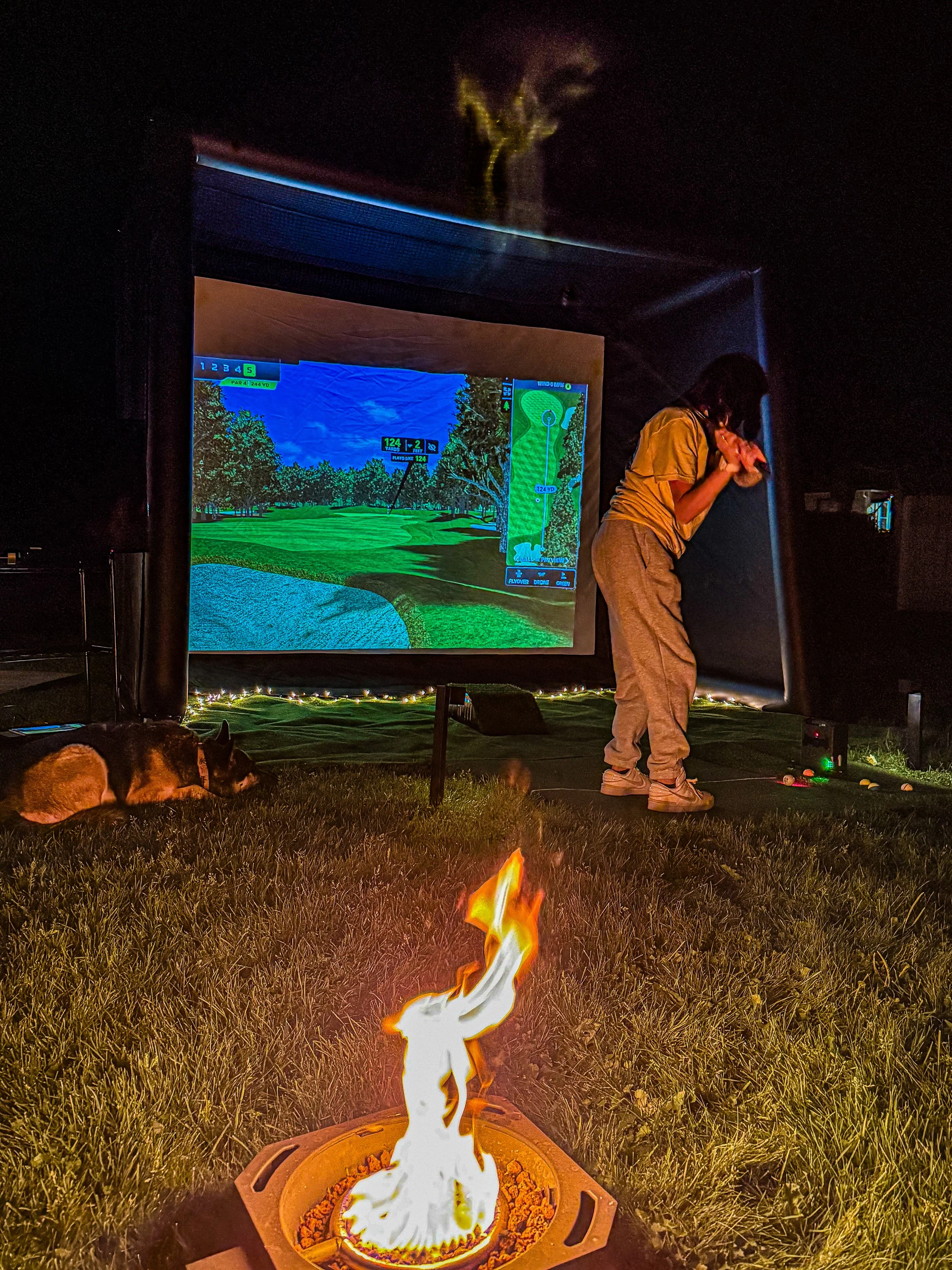 A person standing next to an indoor golf simulator screen at night, with a dog lying on the grass nearby, and a small fire burning in the foreground.